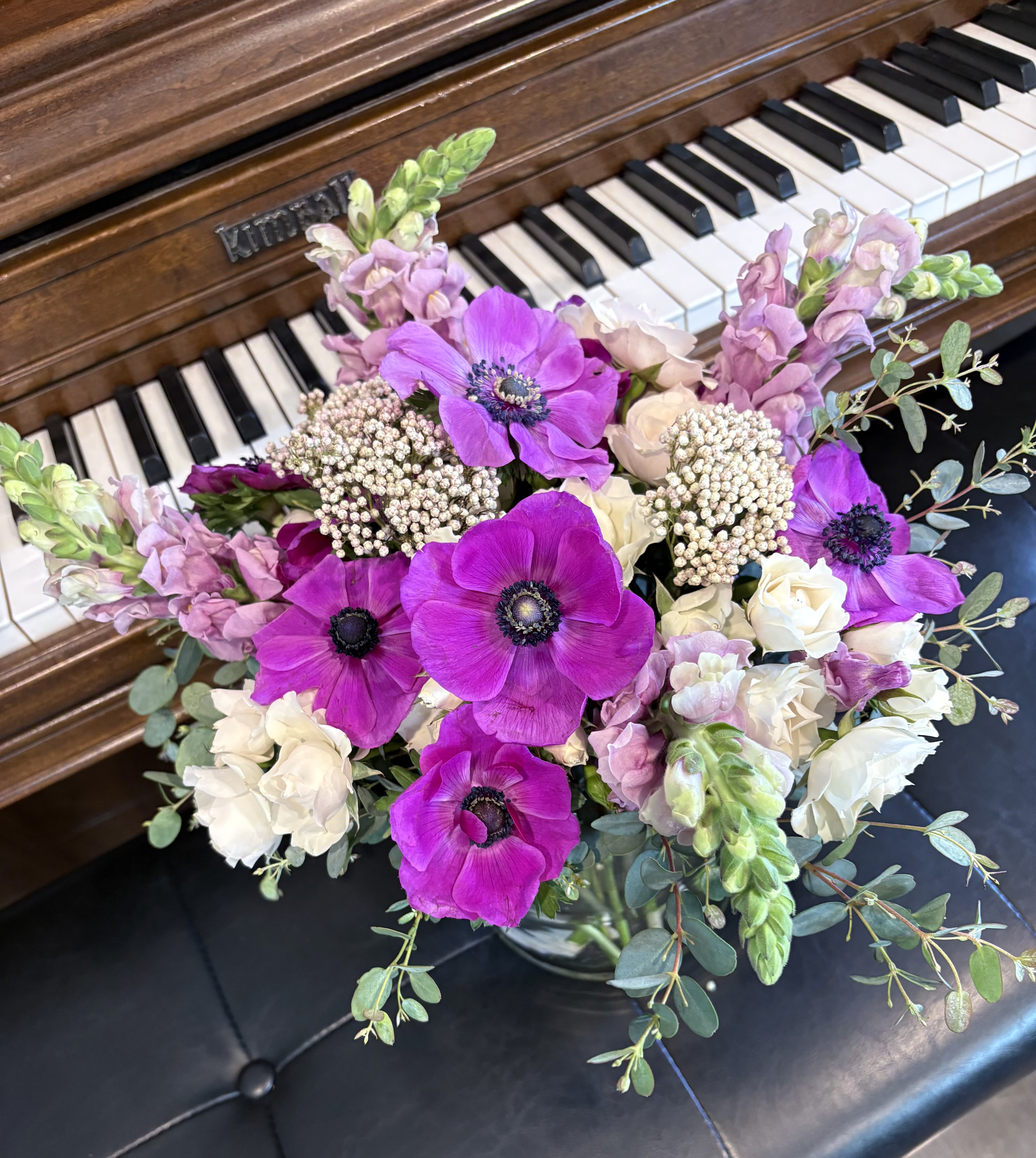 Purple anemones  - Vivid purple anemones take center stage in this floral arrangement, paired with creamy white roses and soft pink snapdragons for a romantic birthday bouquet or anniversary surprise. The lively magenta and blush palette reads both celebratory and gentle, making it a lovely choice for Mother's Day or a thoughtful "just because" gift that will make the person you're thinking of feel seen and cherished.  Arranged in a clear glass vase, the rounded, full bouquet features layered textures-velvety anemone petals, classic rose blooms, and the vertical grace of snapdragons-finished with airy eucalyptus for a loose, natural silhouette. The mix of saturated purple, pale blush, ivory, and fresh green creates eye-catching contrast while feeling elegant on tabletops or entryway consoles. Standard sizing fits most spaces; change water every 2-3 days and trim stems at an angle to keep blooms looking fresh longer.