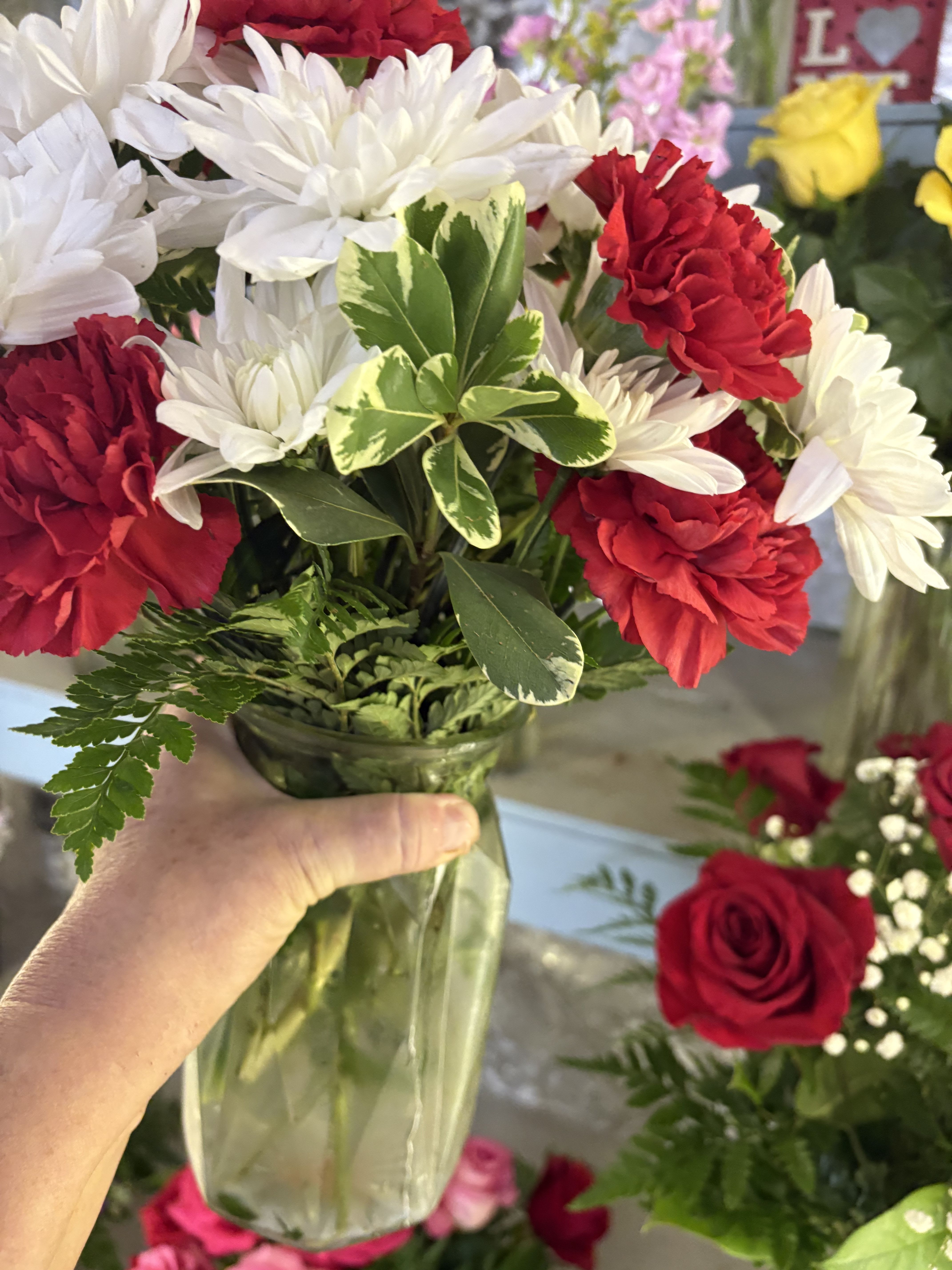 Designers Choice ( Standard, Deluxe and Premium) - Bright red carnations paired with crisp white daisy-like blooms create an energetic, classic look that makes a thoughtful birthday bouquet, a cheerful get-well arrangement, or a warm "just because" surprise for the person you're thinking of. The red-and-white palette reads both festive for holidays like Christmas or Father's Day and friendly enough for everyday moments such as Thank You, Thinking of You, or Congratulations.  Hand-gathered in a clear glass vase, the mixed bouquet is arranged in a loose, rounded shape with mixed greenery and variegated foliage for texture and contrast. Available in Standard, Deluxe, and Premium sizes to suit intimate gestures or larger celebrations, it works beautifully for occasions from graduations, anniversaries, and housewarmings to sympathy and funeral settings. Care is simple: fresh-cut stems in clean water, change the water every other day, and trim stems slightly for longer vase life.