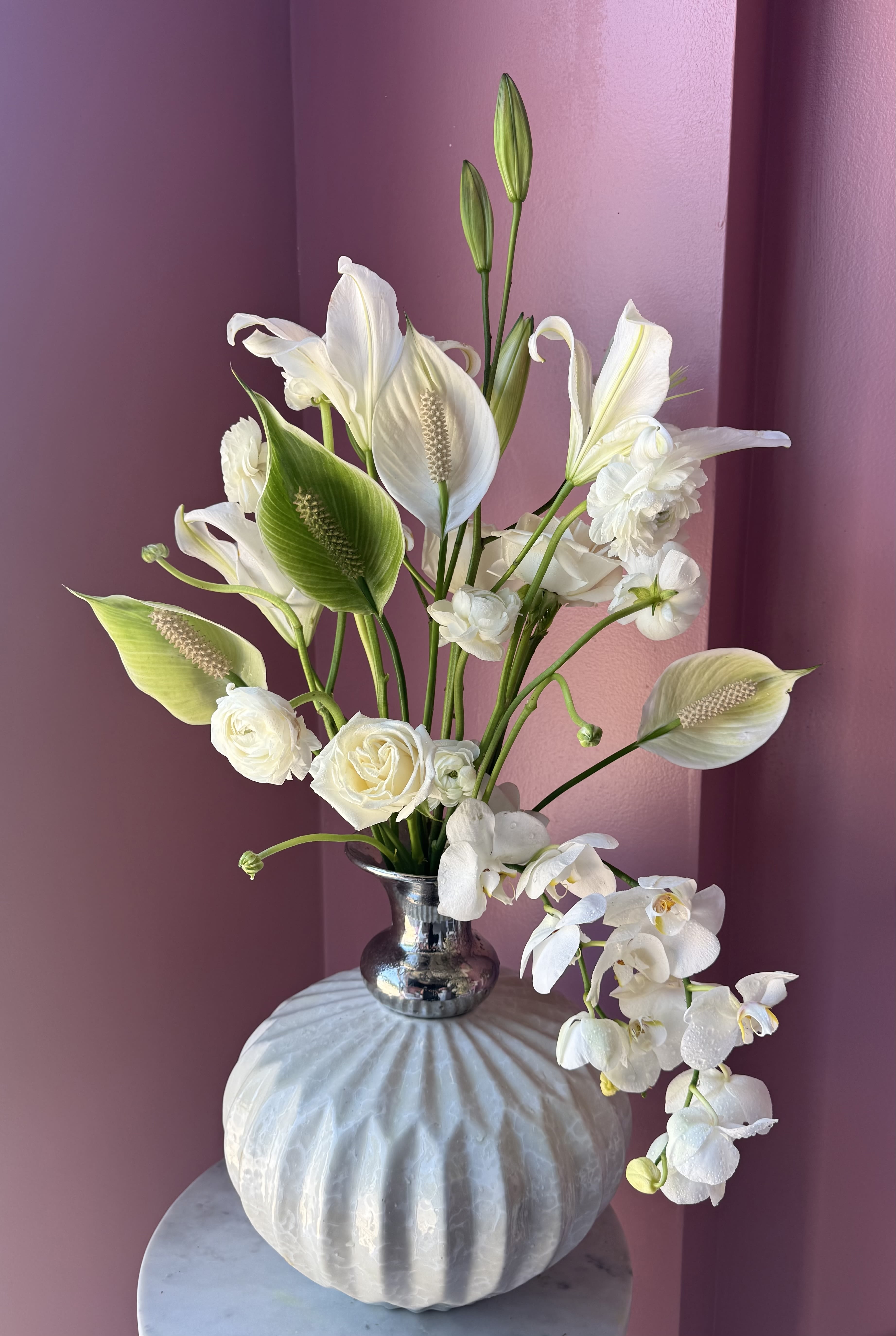 Petals &amp; Polish  - White calla lilies and orchids arc gracefully above cream-toned roses in a tall, airy composition that reads both modern and serene - an elegant flower arrangement for sympathy, a wedding, or a thoughtful thank-you gift. The predominantly white and green palette with subtle purple undertones creates a calming, refined presence that makes the recipient feel quietly seen and comforted.  The blooms are arranged in a reflective silver vase set on a white pumpkin-shaped container for a slightly sculptural, contemporary silhouette. Textures contrast softly: the smooth calla spathes, the delicate orchid blooms, and the classic rose petals combine for visual depth while remaining light and clean. Sized to command a tabletop or entryway without overwhelming a room, this arrangement arrives ready to display and bring a gentle, sophisticated touch to the moment you're marking.