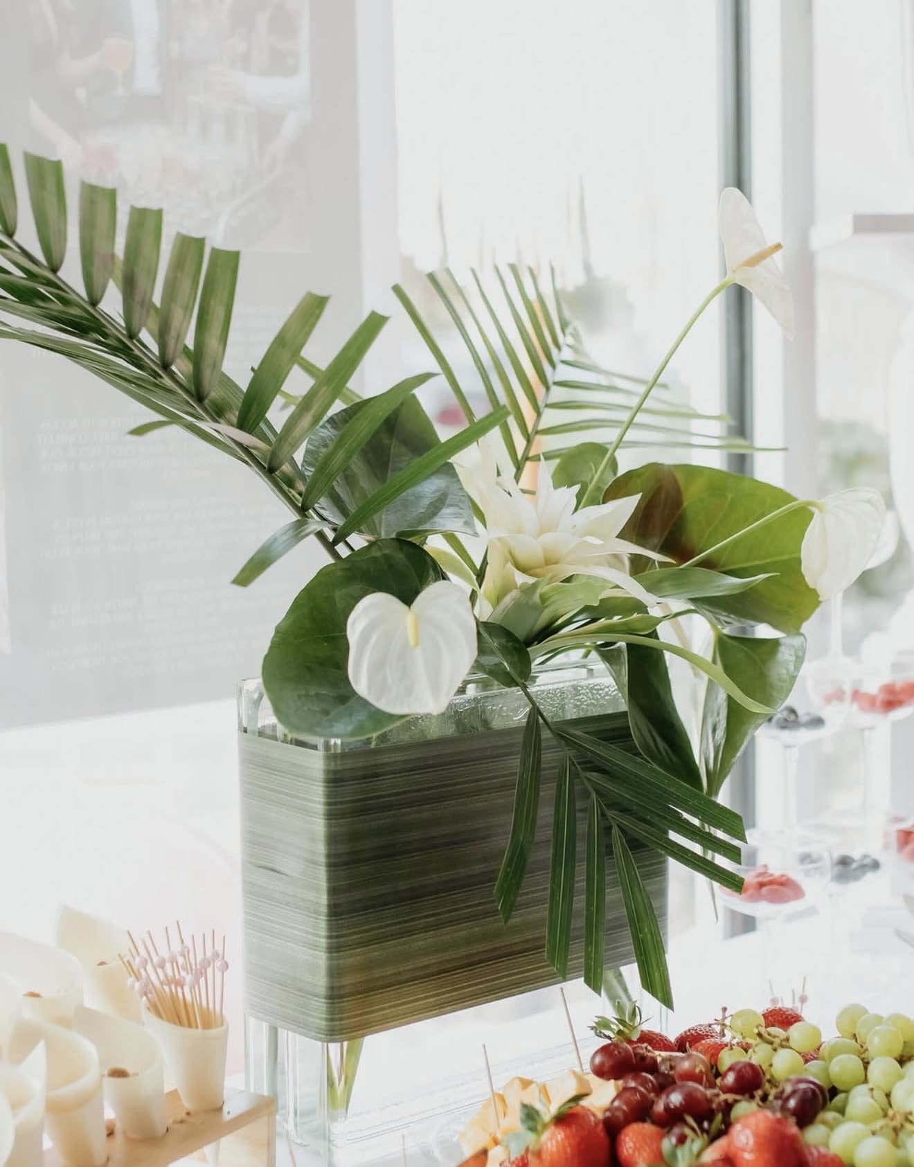Linear Luxury  - Crisp white tropical blooms rise above broad glossy leaves and layered palm fronds in a modern, sculptural arrangement - a sleek flower arrangement perfect for housewarming, everyday decor, or a serene celebratory gift. The vertical lines and airy composition give it a refined, contemporary tropical feel that will make the person you're thinking of feel soothed and seen in a bright, modern space.  Carefully arranged in a square glass vase, the design pairs pure white flowers with lush green foliage for a clean, minimalist palette that complements living rooms, entryways, or office settings. Tall palm fronds and glossy leaves create height and texture while keeping the overall look light and elegant; dimensions and vase size are consistent with a tabletop statement piece. For best freshness, replace vase water every 2-3 days and recut stems at an angle before arranging; ideal as a housewarming gift or a calm, modern accent to mark a quiet celebration.