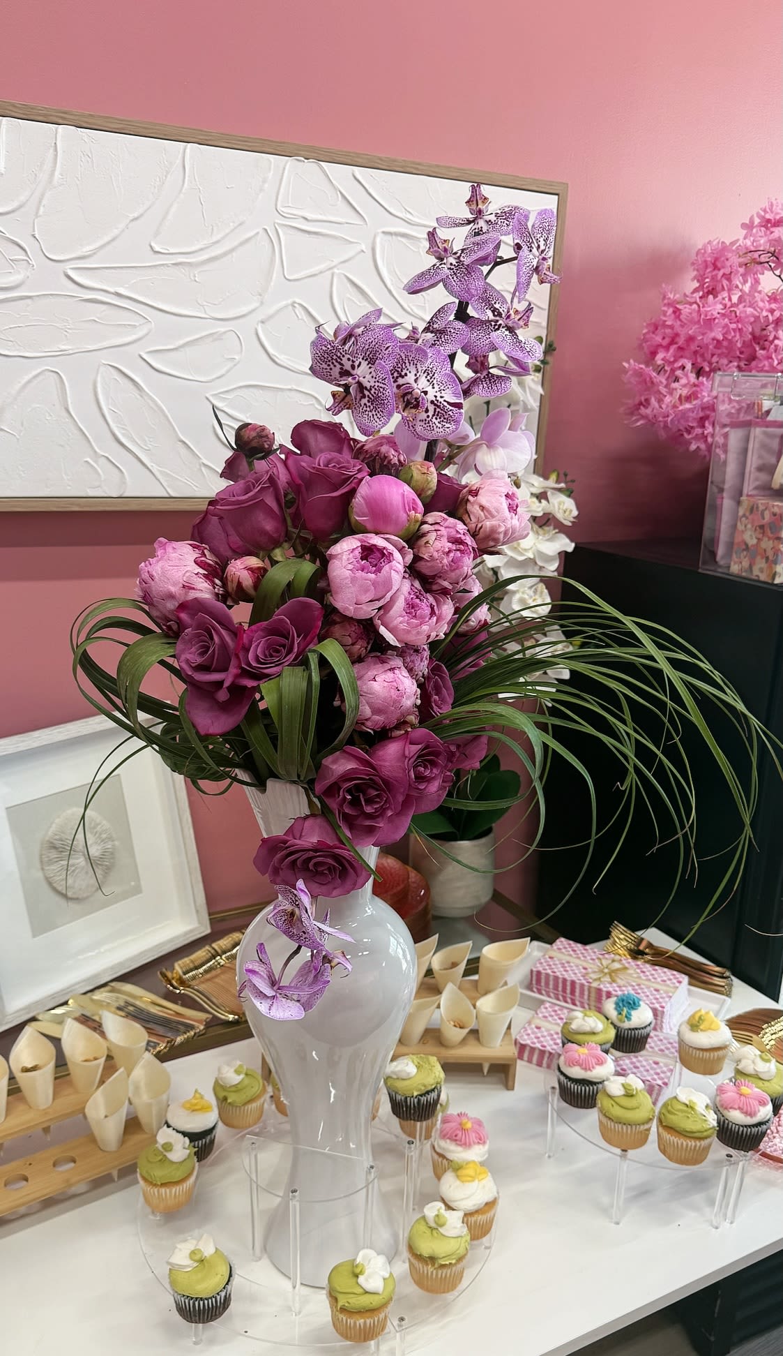 Plum Perfection  - Rich plum roses and spotted purple Phalaenopsis orchids mingle with soft pink peonies in a tall white ceramic vase-an arrangement that reads like celebration. As a birthday bouquet, anniversary gift, or centerpiece for a spring soiree, this flower arrangement makes the person you're thinking of feel seen and celebrated with its feminine, festive palette of purple, pink, white, and fresh green accents.  The design is sculptural: long looping blades of greenery arc outward to give dramatic height and movement while clusters of blooms create a lush, textured core. The white ceramic vase keeps the focus on the deep plum roses, delicate orchid blooms, and ruffled peonies; the styling in the photo with clear acrylic cupcake stands, pink checkered gift boxes, and wooden serving trays shows how well this piece anchors a dessert table or event display.  Perfect for marking birthdays, anniversaries, or spring gatherings, Plum Perfection sends a message of thoughtful, elegant celebration. The tall vase makes it easy to place on a buffet or entry console; fresh-cut flowers arrive ready to display-trim stems and change the water every two days to keep the arrangement looking its best.