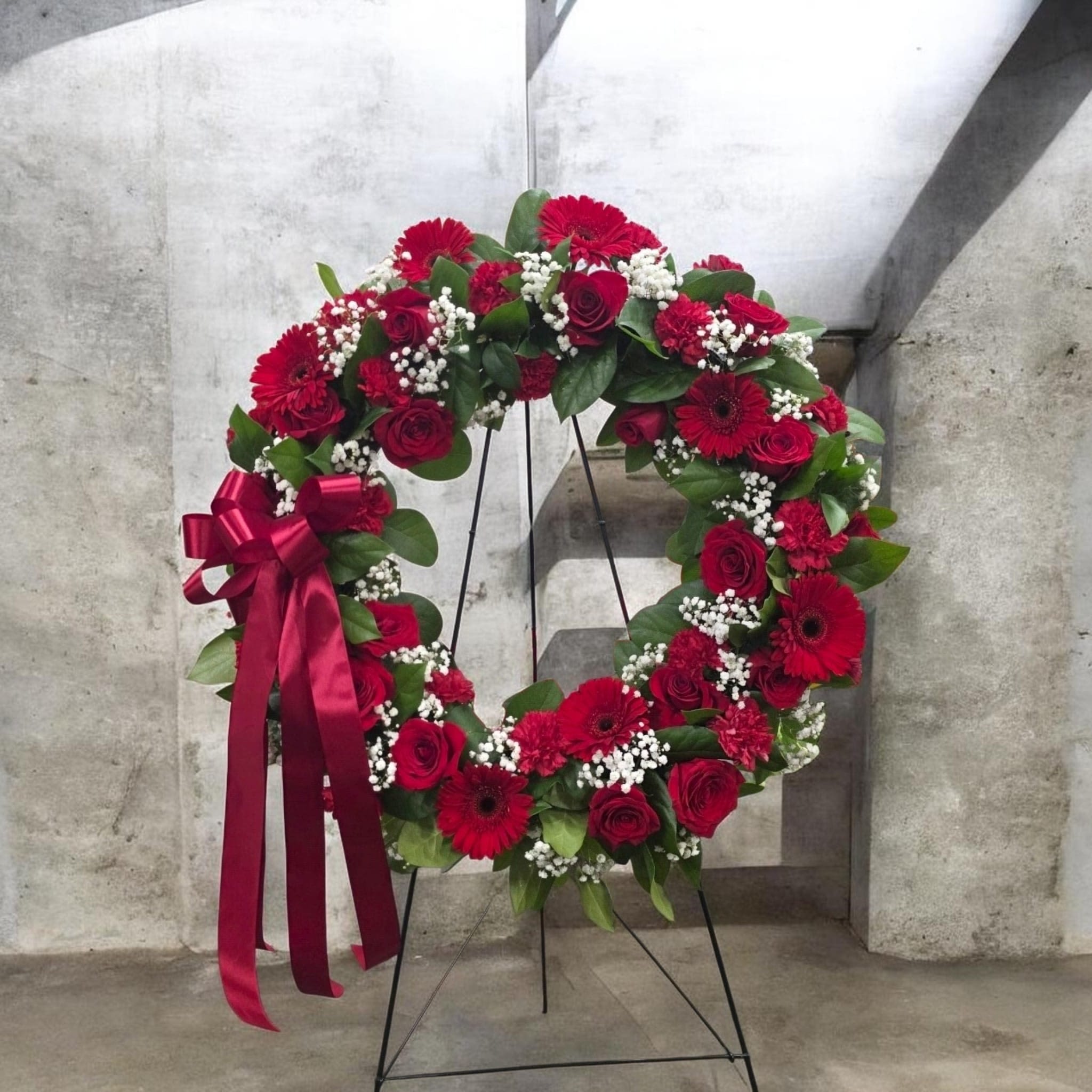 Sacred Grace Floral Tribute - A rich circular wreath of deep red roses, gerbera daisies, and carnations makes a solemn, heartfelt statement-perfect as a sympathy arrangement or memorial floral tribute. The Sacred Grace Floral Tribute is designed for funerals and remembrance services, offering a formal, comforting presence that lets the person you're honoring feel respectfully remembered.