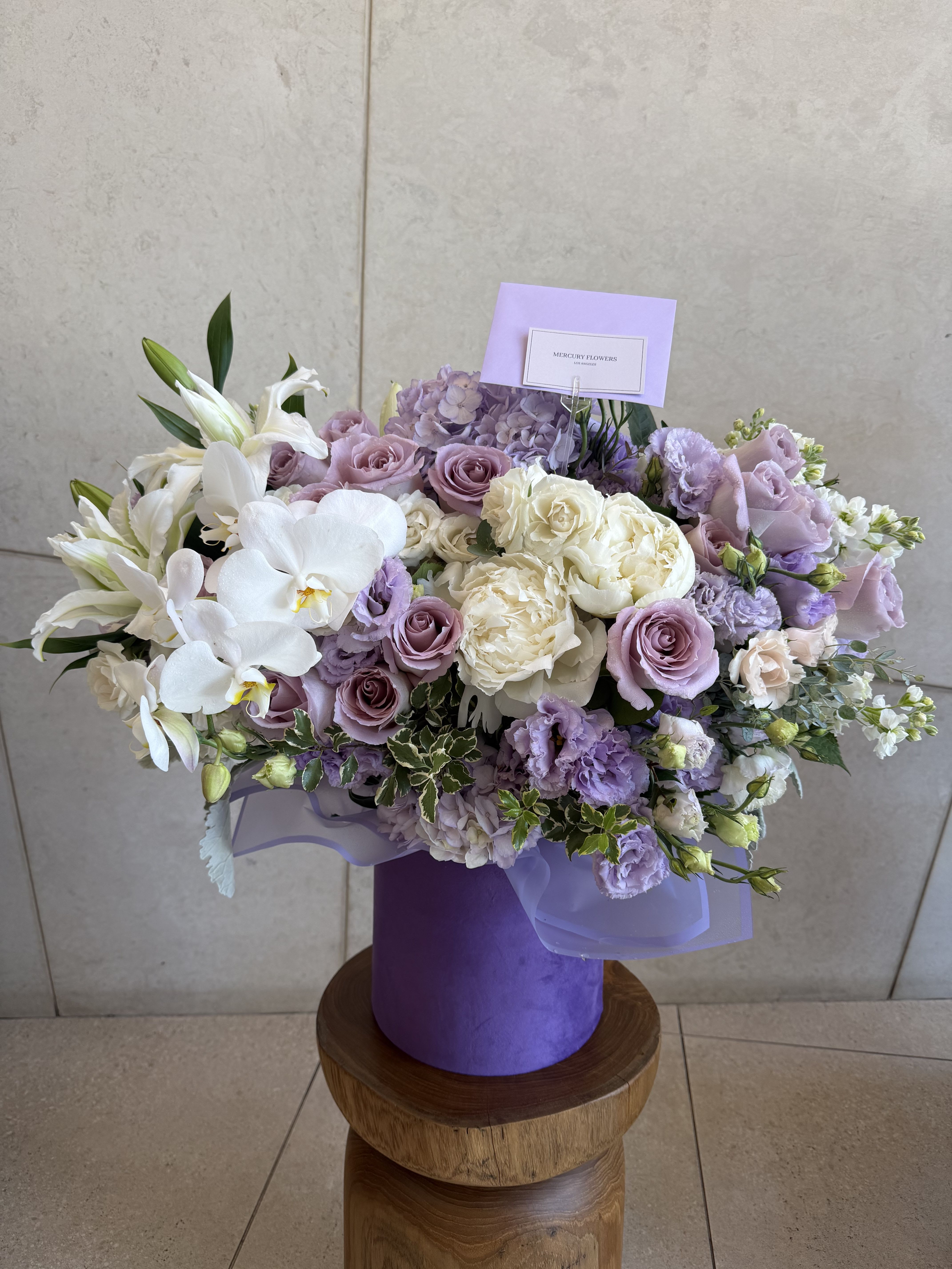 Lavender Dream Orchid Box - A luxurious garden-style flower arrangement featuring premium lavender roses, white premium orchids, fragrant lilies, lush peonies, hydrangeas, lisianthus, and seasonal blooms artfully arranged in our signature lavender velvet box