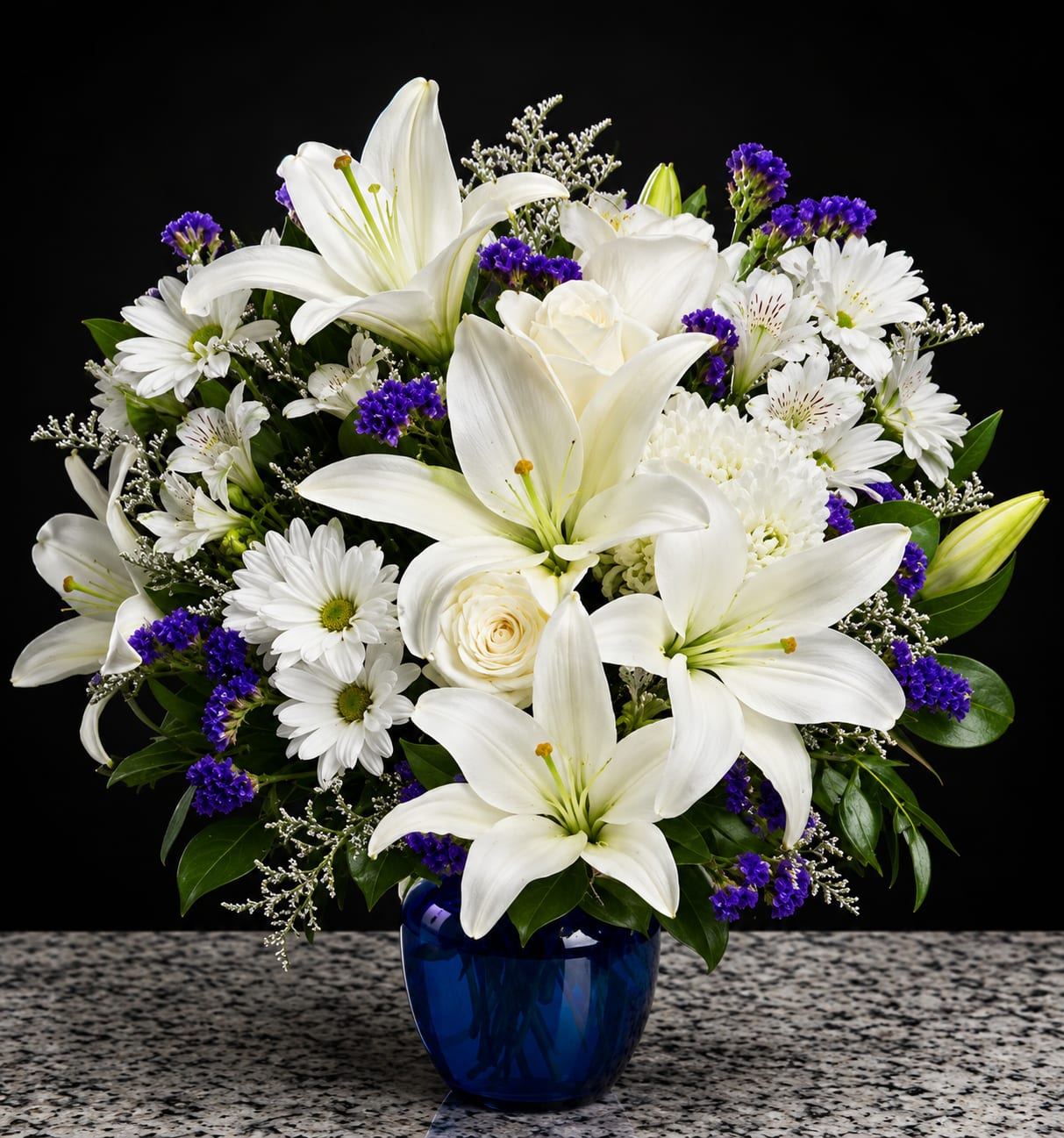 Blue Sapphire Elegance - Big, luminous white lilies take center stage in this elegant flower arrangement, making it an ideal sympathy bouquet or a graceful thank-you gift. The mix of white roses and daisies gives the design a serene, classic feel while the small purple filler blooms add just the right touch of depth-perfect for sending calming, thoughtful sentiments or marking a quiet anniversary.  Set in a cool blue glass vase, the rounded composition balances bright whites against deep purple accents and fresh green foliage for a crisp, formal look. The full, compact shape reads as polished and composed on a mantle or bedside table; the vase height and overall size make it easy to place wherever the person you're thinking of will see it and feel comforted.  This arrangement is meant to convey sympathy, appreciation, or gentle celebration-she'll feel seen and cared for when these blooms arrive. Standard care: keep in fresh water, trim stems every few days, and place away from direct heat to extend vase life.