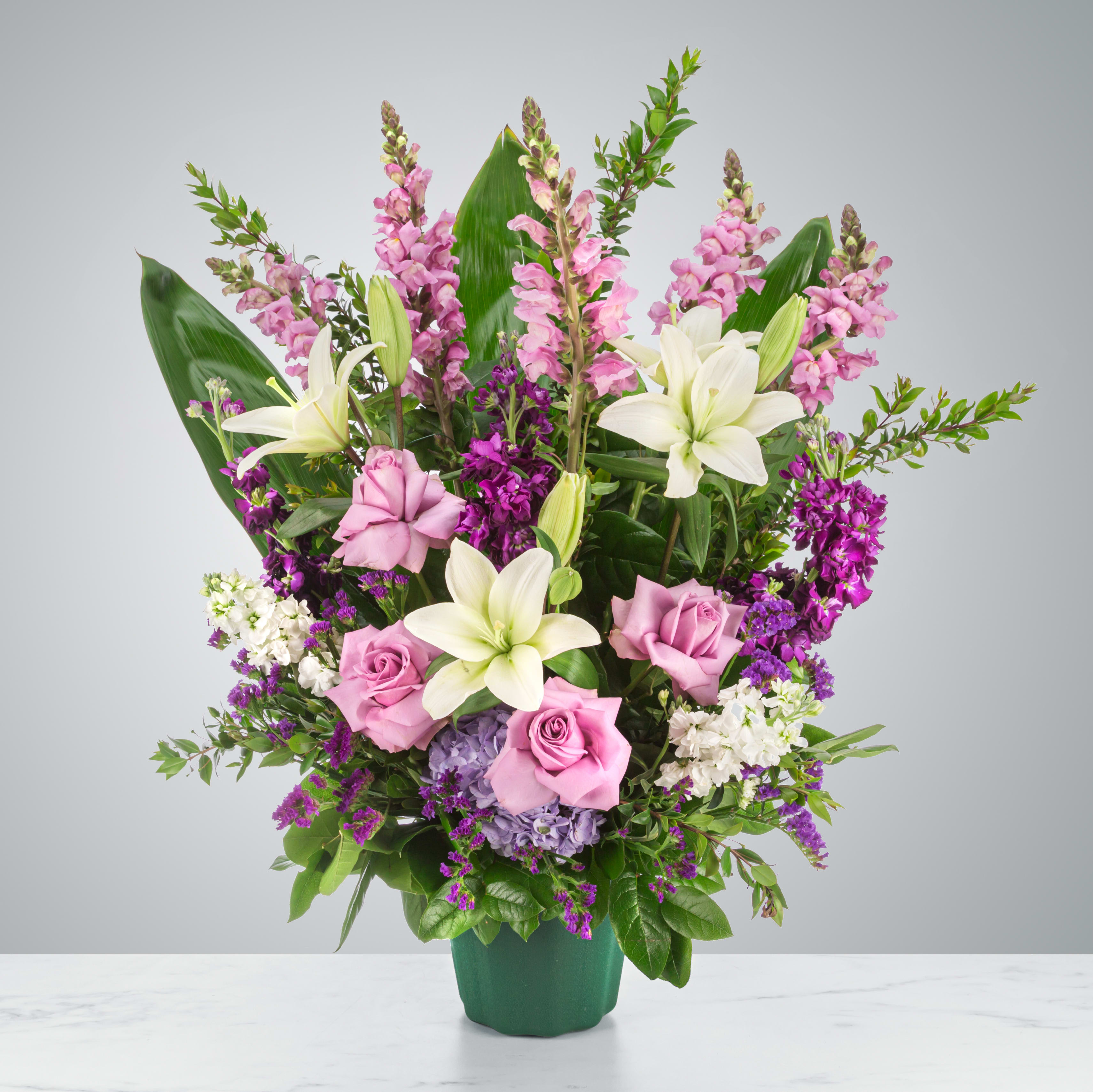 Tall arrangement of white lilies and lavender roses with purple flowers in a green pot