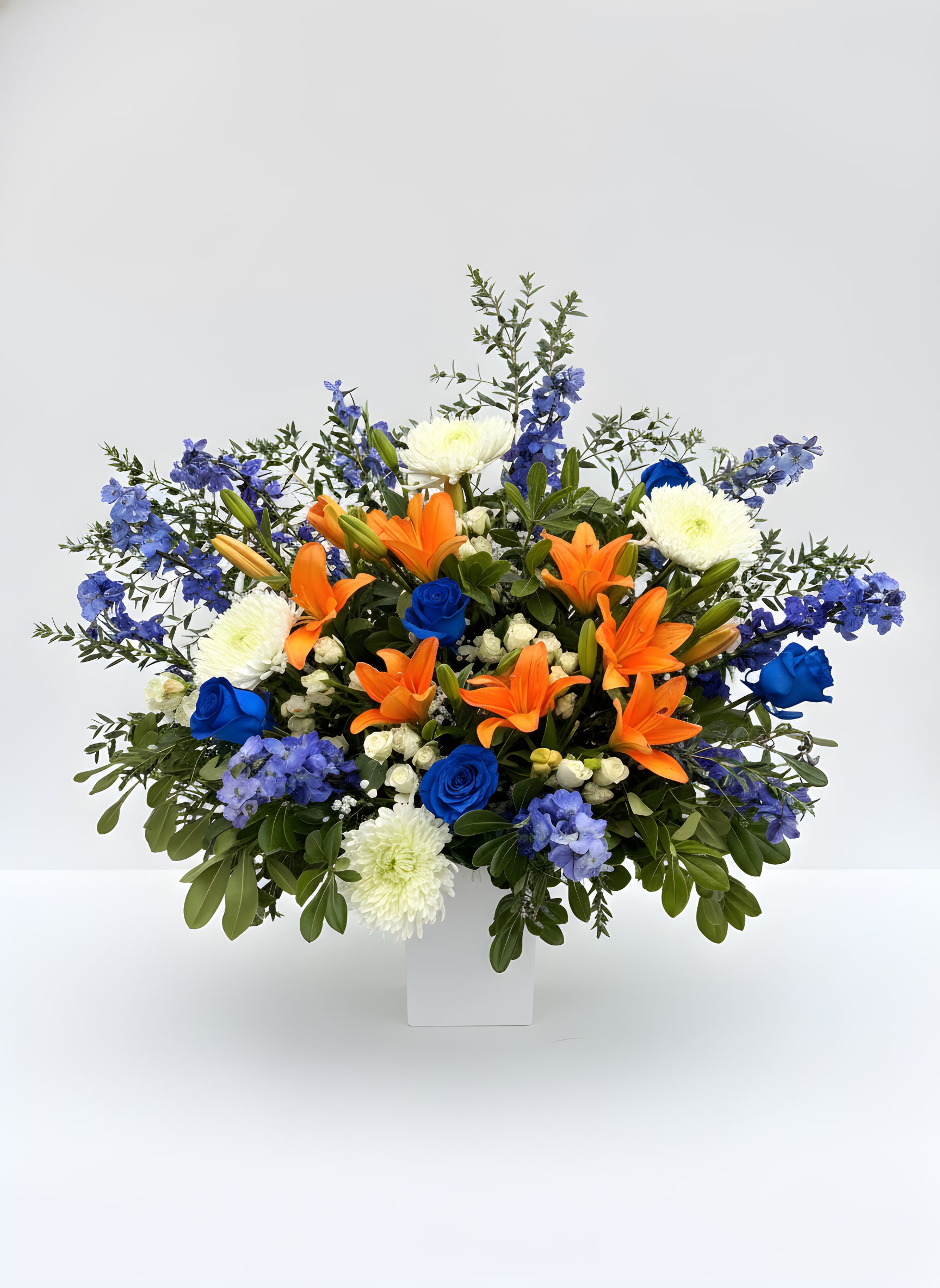 Sunrise Moon - This flower arrangement features lilies, roses, chrysanthemums and airy delphinium arranged to fan outward from a white rectangular vase. The mixed palette of blue, orange and white creates strong visual movement, designed to sit on a mantel, entry table, or celebration spread, it makes an immediate impression without overwhelming the room. 