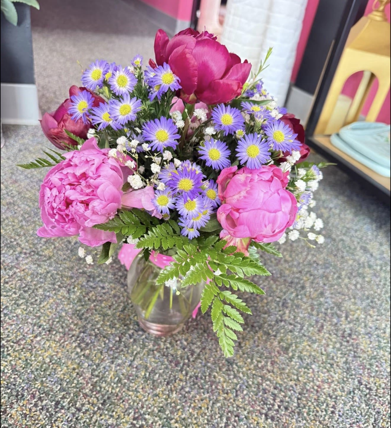 1/2 Dozen Peonies - Big, blousy hot-pink peonies take center stage in this half-dozen peony arrangement, paired with lavender daisy-like asters and delicate white filler for a garden-fresh look perfect for a birthday bouquet, Mother's Day gift, or a cheerful "thank you." The mix of round, velvety peony heads and airy purple accents creates a lively, feminine feel that will make the person you're thinking of feel truly seen and celebrated.  Arranged in a clear glass vase and finished with a playful pink polka-dot ribbon (plus an additional bright pink ribbon at the base), the design reads bright and springlike on any tabletop. Colors include shades of pink, purple, white, and green; textures range from the soft, full peonies to the tiny, crisp filler blooms. Sized as a half-dozen of peonies, it arrives ready to display-refresh the water every few days and recut stems for longer enjoyment.