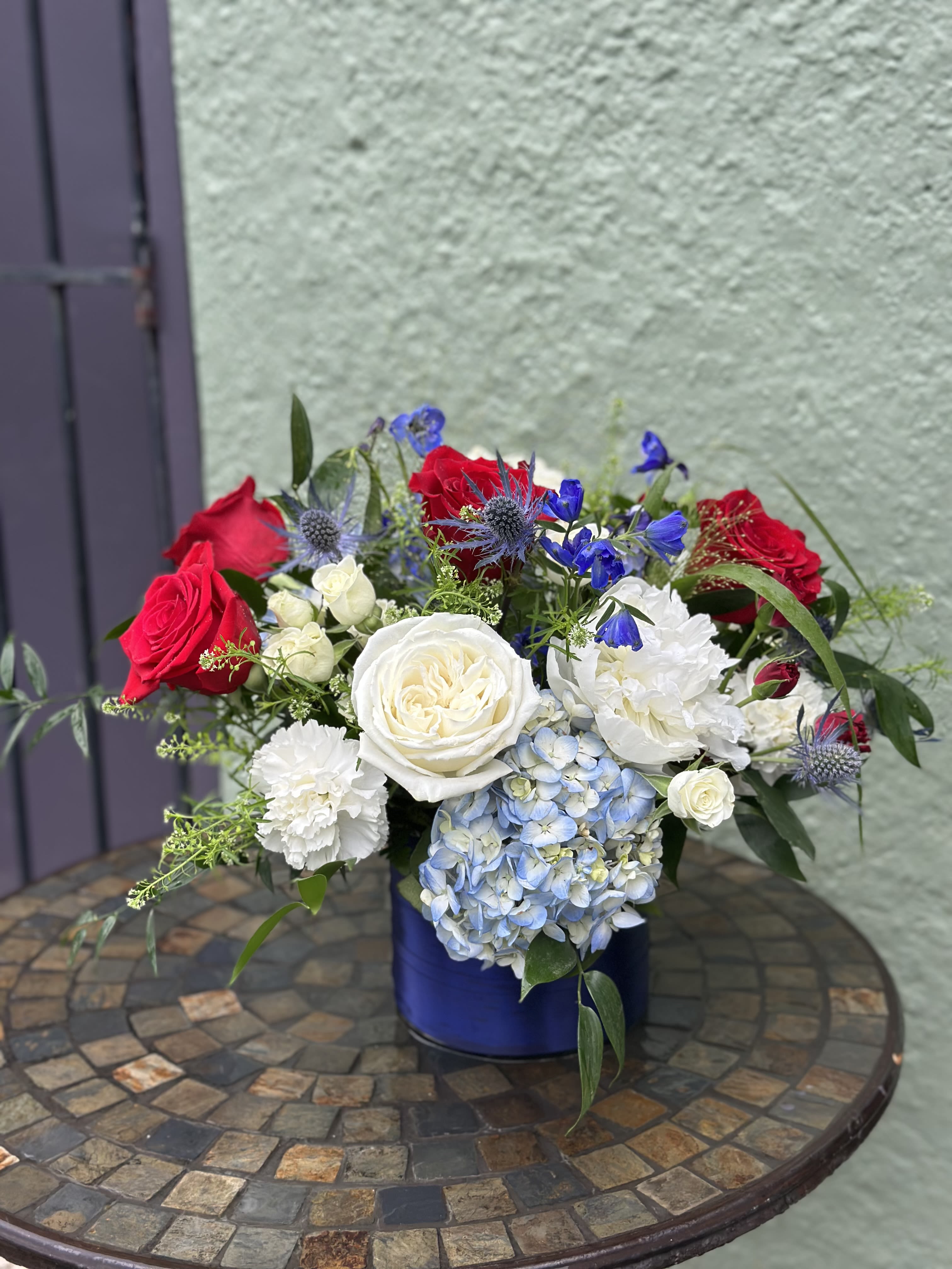 Americana - A bold red-white-blue centerpiece that reads both patriotic and celebratory, this flower arrangement is perfect for your Fourth of July gathering or block party. The mix of red and white roses, pale blue hydrangeas and small blue accent blooms create a contemporary, eye-catching Americana look. The rounded, full shape and balanced color blocking make this arrangement easy to place on a dining table, coffee table or as a striking focal point at your gathering.