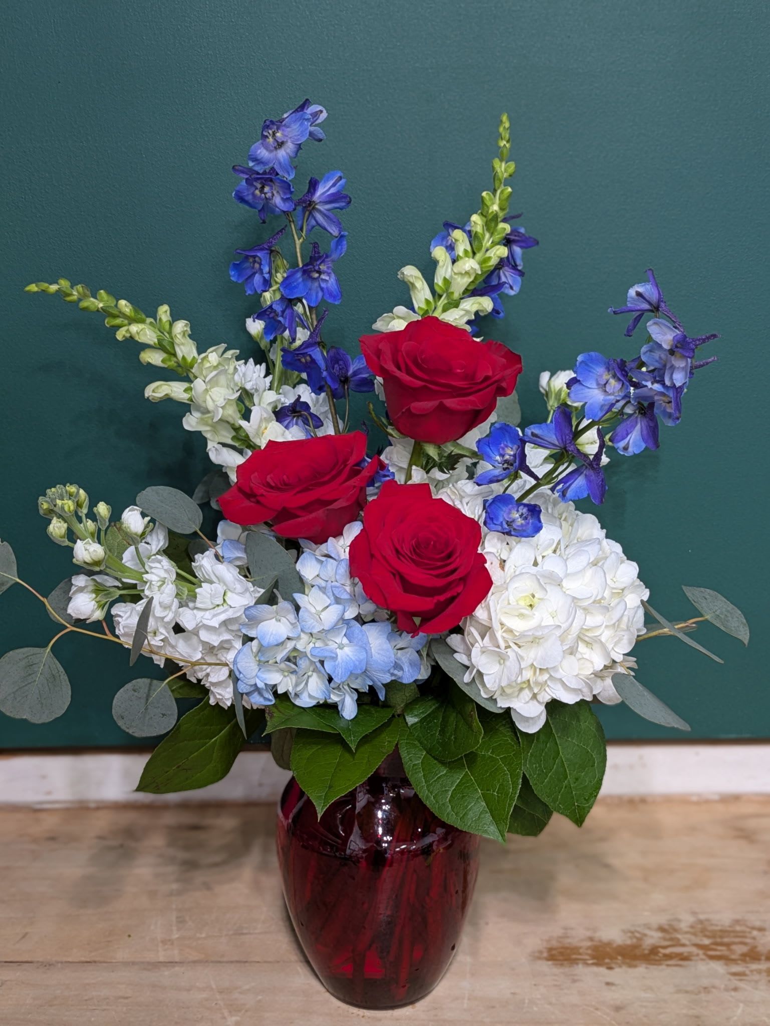 Viva America - Limited Time - Happy 4th of July! Three bold red roses stand out against clouds of white and pale blue hydrangea and tall spikes of delphinium for a patriotic, celebratory flower arrangement perfect for birthdays, anniversaries, or Fourth of July gatherings. The red, white, and blue palette-lifted by touches of purple delphinium and soft gray-green eucalyptus-creates a vivid, romantic impression that will make the person you're thinking of feel honored and seen.  This rounded, full composition pairs the dense texture of hydrangea with airy height from delphinium and the sweet, columnar presence of white stock. Available in versions with three or five red roses, the arrangement is presented in a deep burgundy glass vase that adds weight and a formal, rich note to the overall look. Dimensions and exact stem counts vary by size selection; eucalyptus and seasonal filler round out the mix to keep the bouquet lush and full.  Ideal for patriotic events, milestone moments, or as a thoughtful thank-you, Viva America reads as both festive and timeless on a mantel, dining table, or office desk. Choose your size at checkout for delivery, and enjoy simple care-fresh water in the vase and a quick trim of stems every few days will help maintain the arrangement's color and shape.