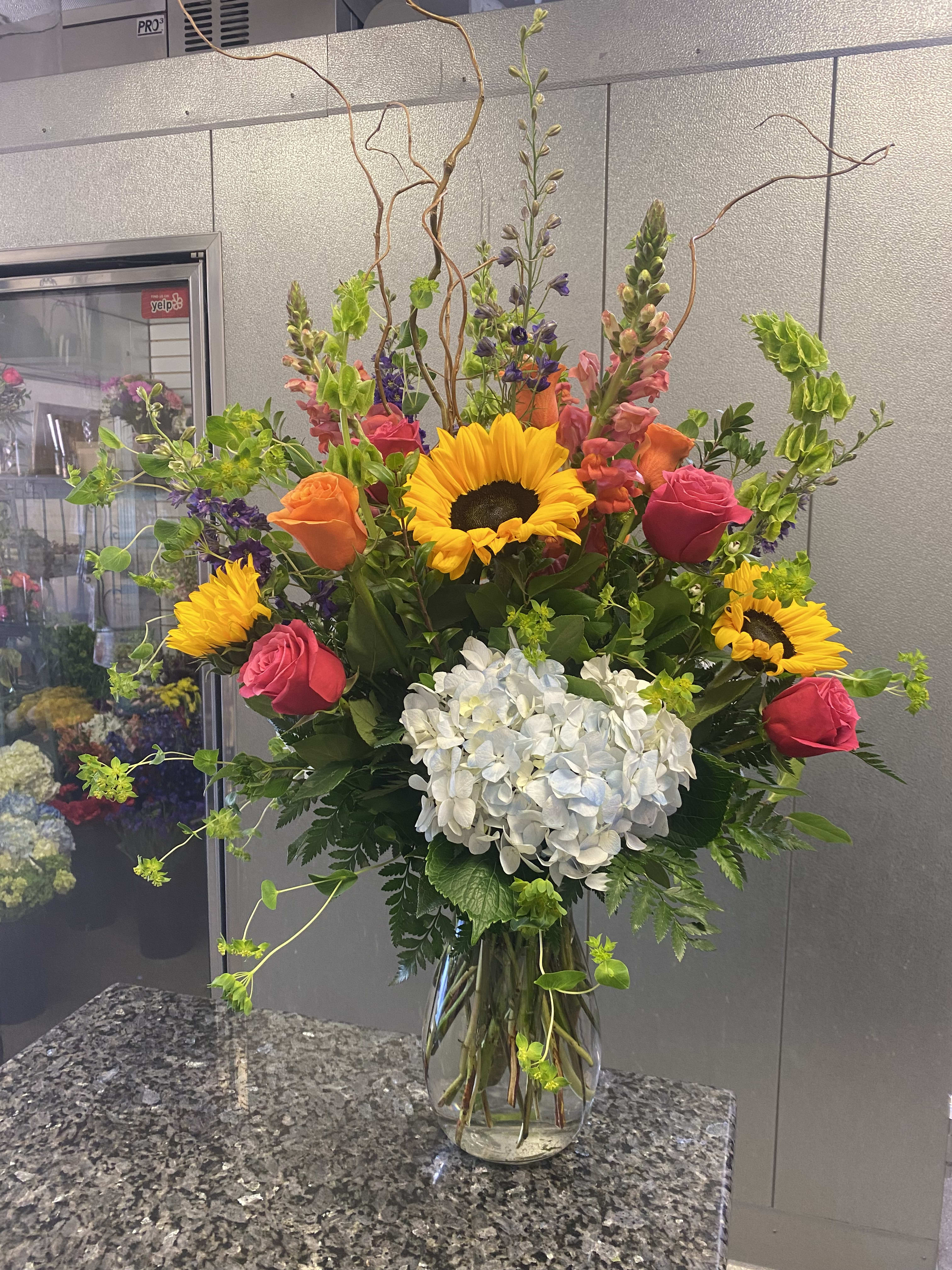 Tall mixed bouquet with sunflowers, roses, and white hydrangea in a glass vase