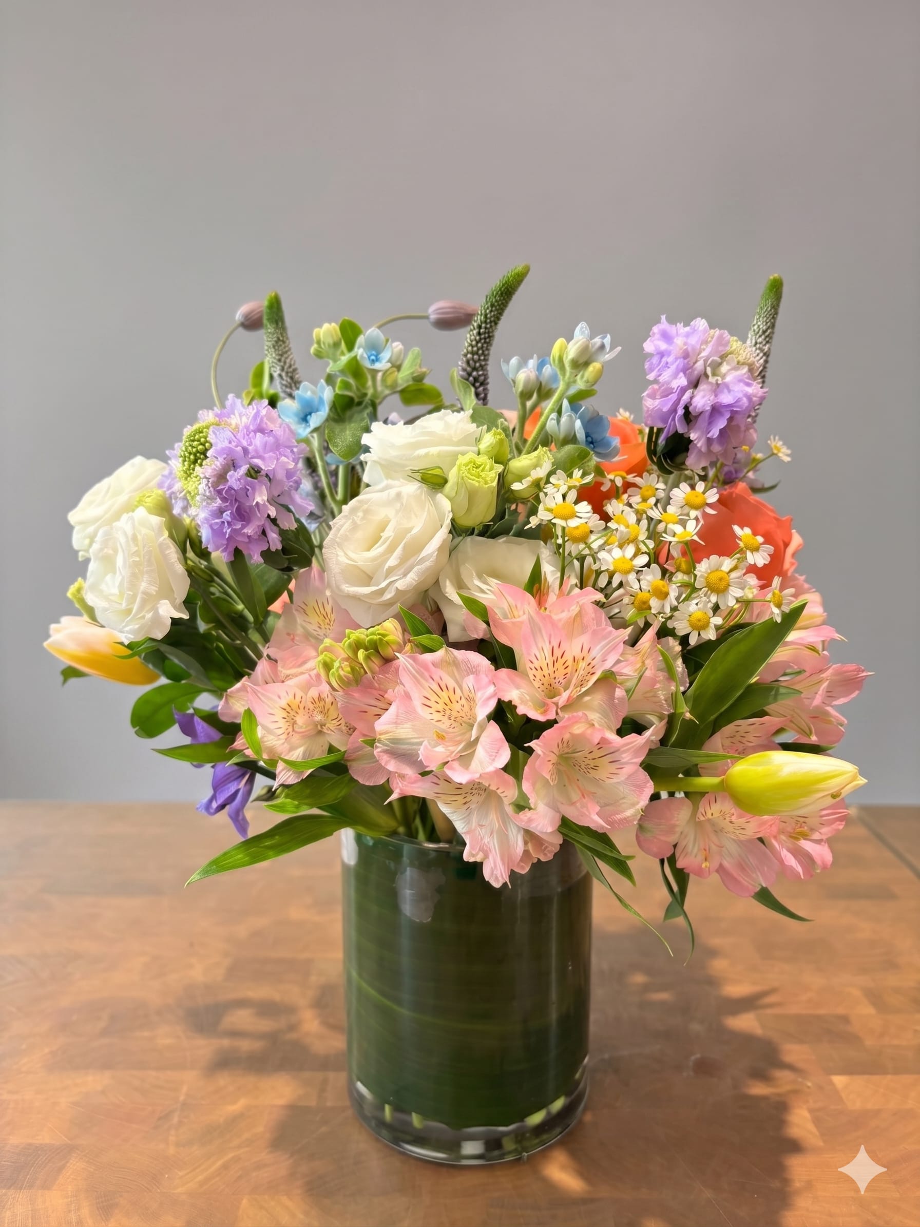 BP3 - Garden Mix Arrangement - A lush garden mix featuring pink alstroemeria, white lisianthus, and purple scabiosa. Accented with yellow tulips, tiny daisies, and set in a clear glass vase with a leaf lining.  Note on Seasonality: The arrangement shown features Tulips (available spring).