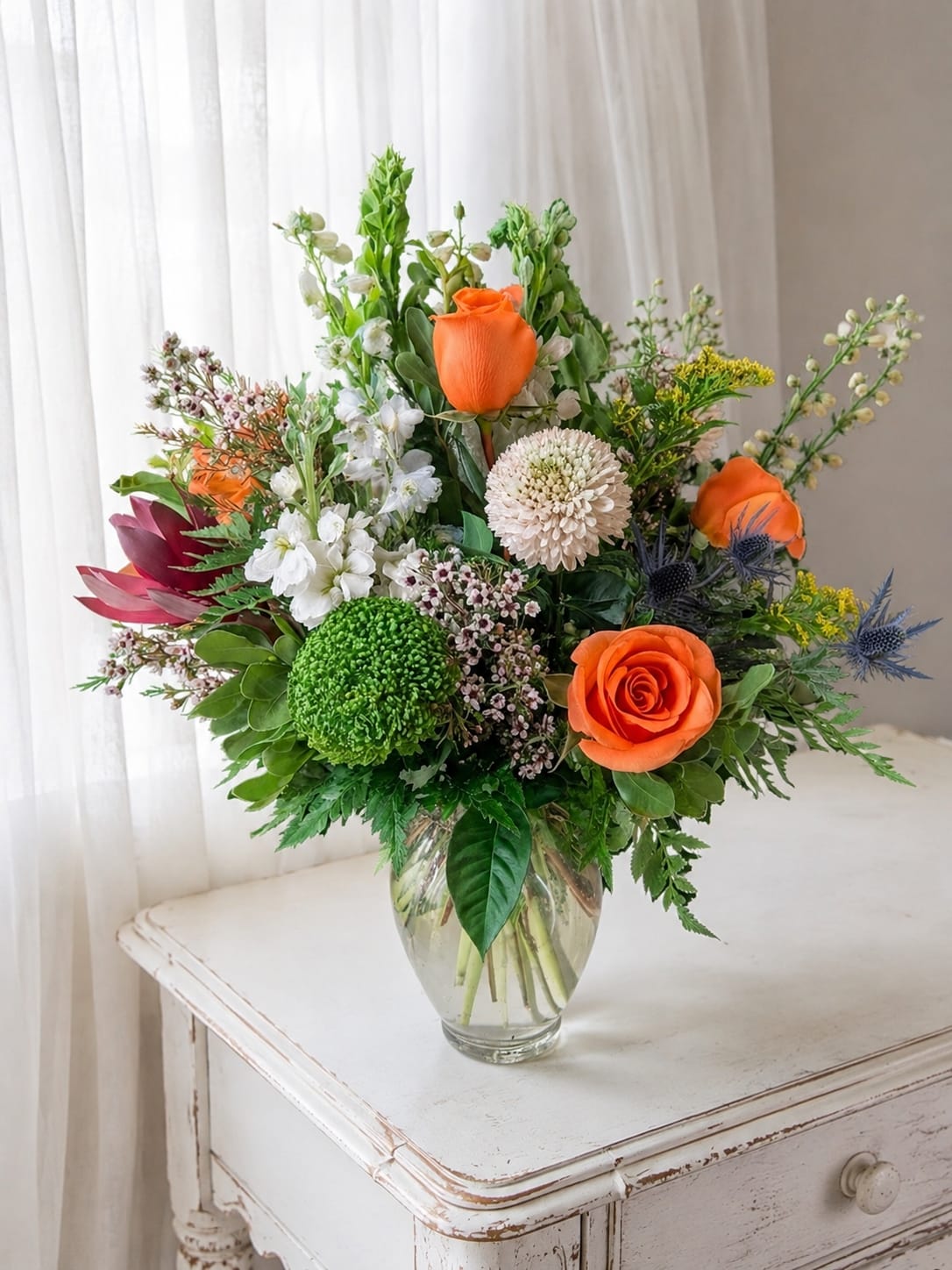 Orange delight - Bright, joyful orange roses take center stage in this birthday bouquet and spring celebration arrangement, designed to make the person you're thinking of feel genuinely celebrated. Orange Delight pairs those saturated roses with clusters of white blooms and a round green chrysanthemum for a vivid, uplifting color story; touches of pink, yellow and soft blue in the airy accents add a playful, garden‑fresh energy that reads cheerful and lively.  The design is full and slightly asymmetrical, with tall snapdragon stems rising above a dense base of roses, daisies and mixed filler for layered texture and movement. Arranged in a clear glass vase, the composition keeps the look clean and modern while letting the bright orange and fresh greens pop. The mix of flower types creates varied shapes - round chrysanthemums, classic rose blooms, spiky snapdragons and simple daisies - for an eye‑catching display from every angle.  Perfect as a thank‑you gift, birthday bouquet, or a sunny pick‑me‑up for someone who loves warm color, this arrangement feels polished yet natural - the sort of bouquet that makes a recipient smile the moment it arrives. Standard vase sizing fits most tabletops; to keep blooms looking their best, change water every two days, recut stems at an angle, and remove any leaves below the waterline.   If any design elements are of major importance to your order, please include them in the florist instructions at checkout or contact us to ensure availability.