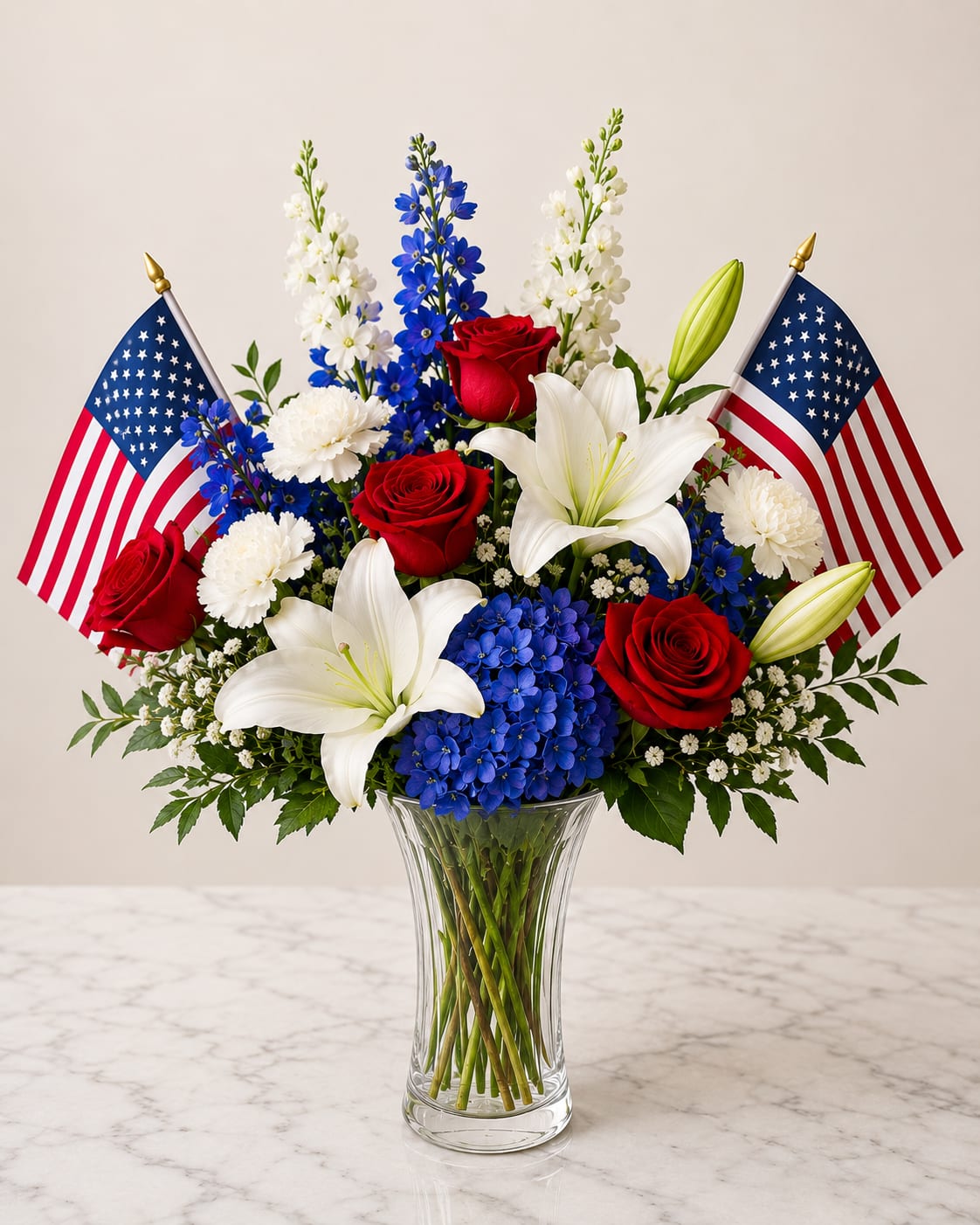 Spirit of America - Bold red roses, white lilies, and sky-blue hydrangeas come together in a patriotic flower arrangement perfect for Memorial Day, Fourth of July, or to honor a veteran. This red, white, and blue bouquet makes a formal, ceremonial statement-the kind of tribute that helps the person you're thinking of feel proudly remembered.  The design pairs lush red roses, fragrant white lilies, blue hydrangea clusters, white chrysanthemums and other white accent blooms in a clear glass vase. Two small American flags rise behind the blooms, adding height and a respectful, celebratory touch. The clean glass vase keeps the focus on the strong color contrast and classic shapes while offering easy placement on mantels, memorial tables, or as a centerpiece.  Choose this arrangement to mark patriotic holidays, military milestones, or to express sympathy with dignity; it reads both celebratory and reverent. Vase size and stem lengths are arranged for a formal presentation suitable for tabletops and altars-simply keep water fresh and trim stems every few days to prolong bloom life.