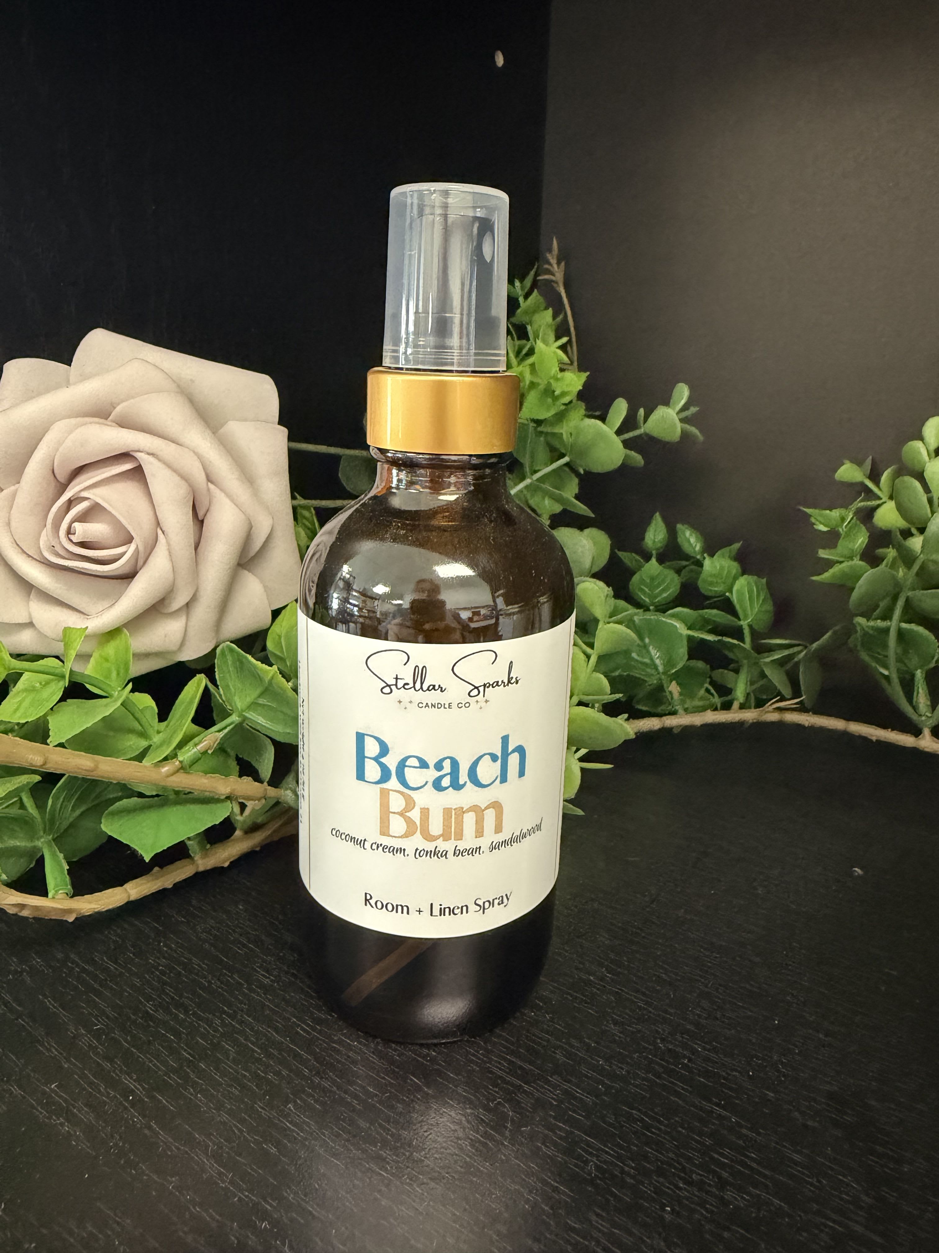 Beach Bum Room Spray - A calming, spa-like room spray that reads like a seaside bouquet-perfect as a small gift or hostess present. This Beach Bum room spray pairs the soothing visual of a large beige rose and soft greenery with a subtle scent meant for bedrooms, bathrooms, or linen closets; it's an ideal pick when you want to make someone feel relaxed and cared for. Use it for a housewarming, thank-you, or just-because moment when you want the person you're thinking of to unwind.  The product features an amber spray bottle with a clear cap and a cream label reading "Beach Bum," styled alongside a neutral beige rose and fresh green foliage in the imagery to suggest gentle, botanical fragrance notes. Colors in the presentation include soft green, white, pink accents and natural beige; the included spray bottle is ready to mist linens, curtains, or the air for an immediate lift. Packaged as a thoughtful little home fragrance, it conveys a quiet, restorative vibe without overpowering the room.  Give this as a thoughtful add-on to a floral gift or on its own to mark a calming occasion-the kind of present that tells someone you want them to relax. For best results, mist lightly on linens and in the air; keep the cap on between uses to preserve the scent.