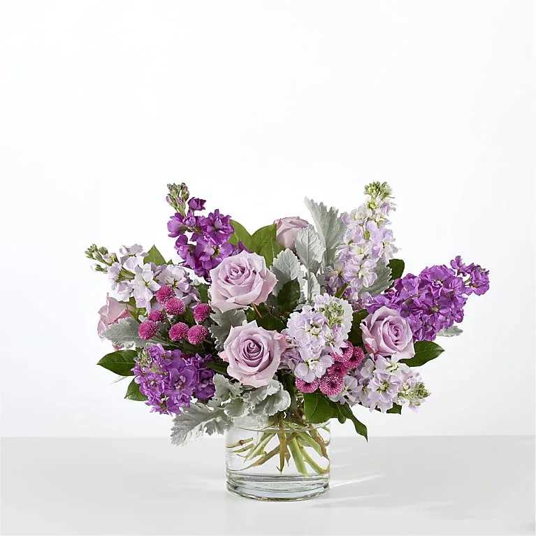 Lilac Dreams - Clusters of lavender roses sit among tall purple and lavender stock, accented by silvery-gray foliage for contrast and texture. Arranged in a clear glass vase the composition spreads outward in a full, rounded silhouette with visible stems for a fresh, natural look. Colors include lavender, purple, pink, white and green; the arrangement reads romantic and refined without being fussy.  Ideal for anniversaries, birthdays, Mother's Day, or as a sympathy bouquet that comforts without volume, this design arrives ready to display in the included clear cylinder vase. Its garden-style shape keeps blooms feeling loose and lively-refresh water every two days and trim stems if desired to prolong vase life.