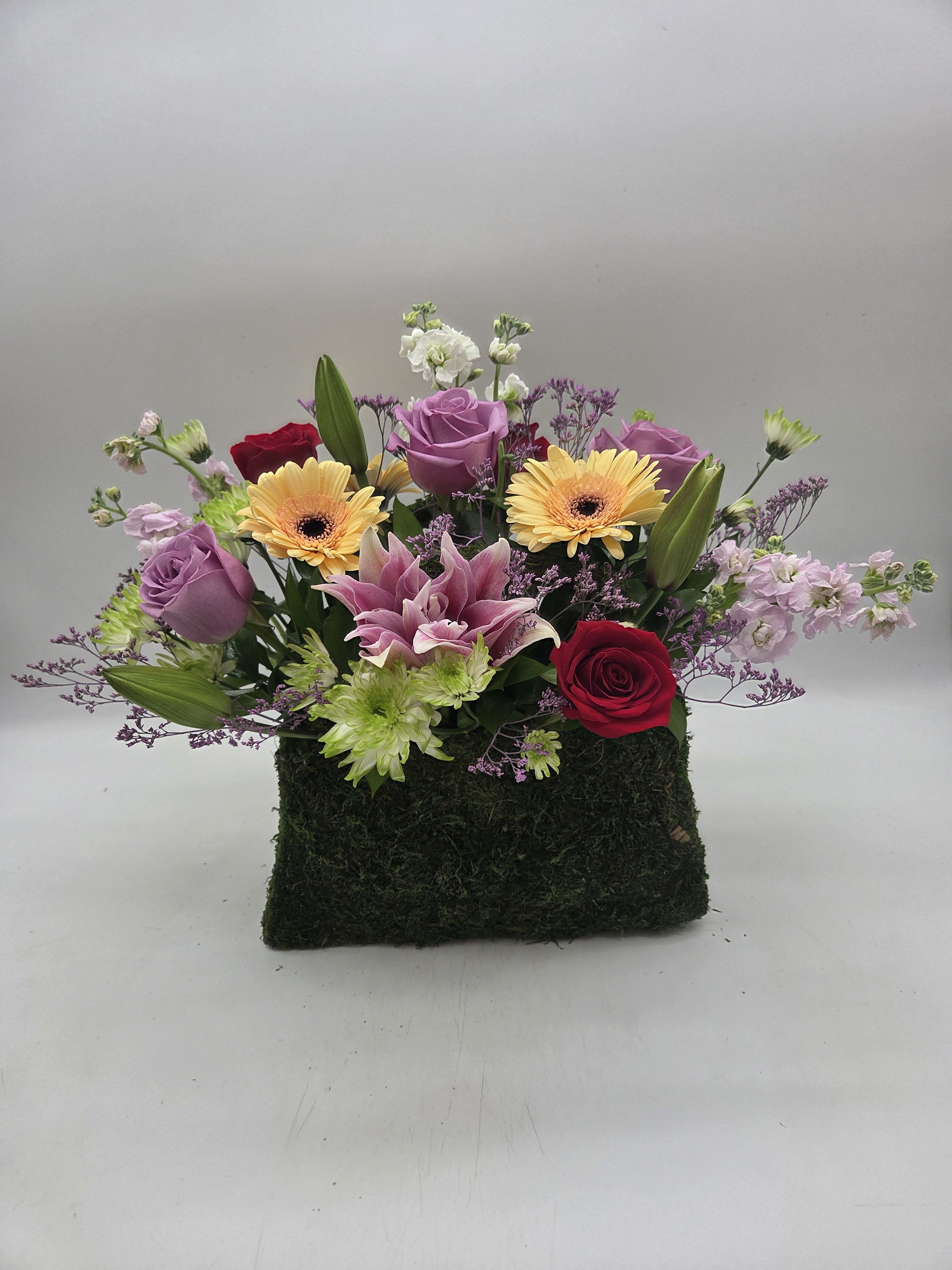Floral Fashionista - Charming floral arrangement designed in a moss-covered purse container, filled with fresh seasonal blooms and lush greenery. This whimsical design brings together the beauty of a garden bouquet with the elegance of a fashionable handbag, making it a perfect gift for birthdays, Mother's Day, get-well wishes, or just because.