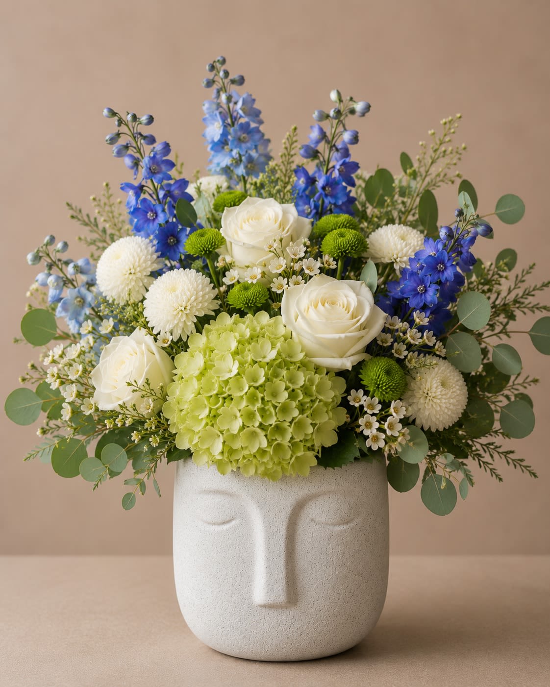 Serenity Bloom Face Planter - Celebrate Dad with a design as timeless and dependable as he is. Dad’s Blue Horizon features striking blue delphinium, elegant white roses, fresh green hydrangea, white button poms, green button poms, delicate wax flower, and eucalyptus artfully arranged in a modern ceramic face vase.  The calming shades of blue, white, and green create a sophisticated and masculine design that symbolizes loyalty, strength, wisdom, and appreciation. Whether you’re honoring a father, grandfather, husband, or father figure, this arrangement is a thoughtful way to express gratitude and love on Father’s Day.  Handcrafted with premium blooms and lush textures, Dad’s Blue Horizon is a memorable gift that brings beauty and meaning to the celebration.