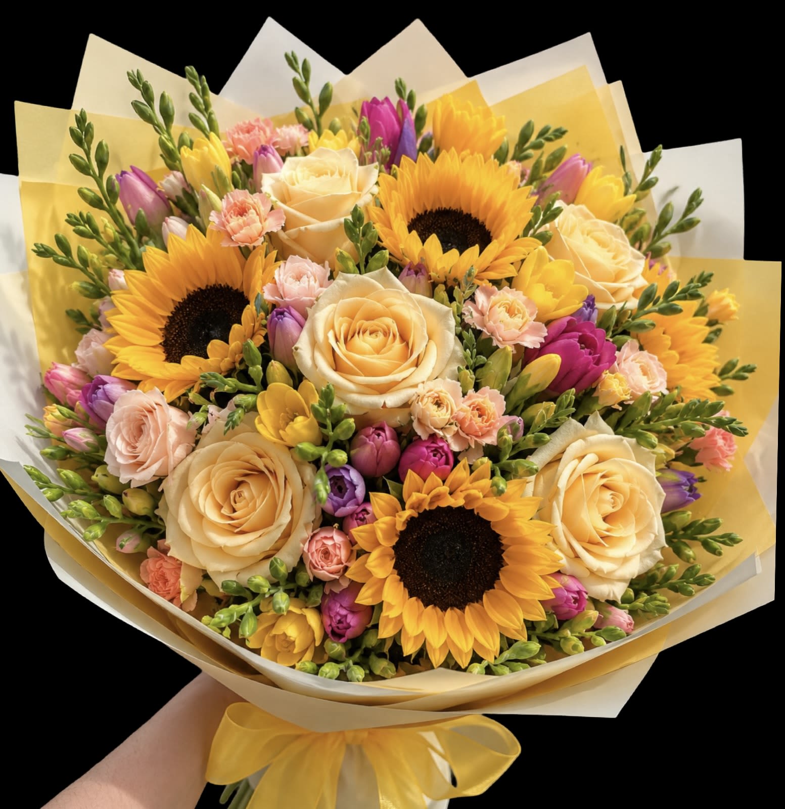 Bountiful Garden Wrapped Bouquet (No Vase) - Golden sunflowers and creamy roses anchor this Bountiful Garden wrapped bouquet for birthdays, get-well wishes, or a bright "thank you" moment. The hand-tied, rounded bouquet reads like a sunlit cottage patch - bold sunflower faces surrounded by soft peach, pink, and purple blooms that make the recipient feel genuinely seen and uplifted.  The mix includes hydrangeas, roses and spray roses, stock, sunflowers, carnations, tulips, snapdragons, and freesia in a lively palette of white, peach, yellow, pink, and purple. It's wrapped in layered yellow and white paper and finished with a yellow satin ribbon for a polished, gift-ready presentation. Dense and abundant with texture - from the sunflower's golden disks to the velvety rose petals and feathery stock - it arrives ready to display or be placed in a vase at home.  Perfect for spring celebrations, Easter or Passover tables, a simple apology, or a Valentine's note that leans cheerful rather than formal, this bouquet is designed to brighten recovery rooms and kitchen counters alike. Size and care: hand-tied, no vase included - trim stems and place in fresh water with flower food to extend the life of the blooms.