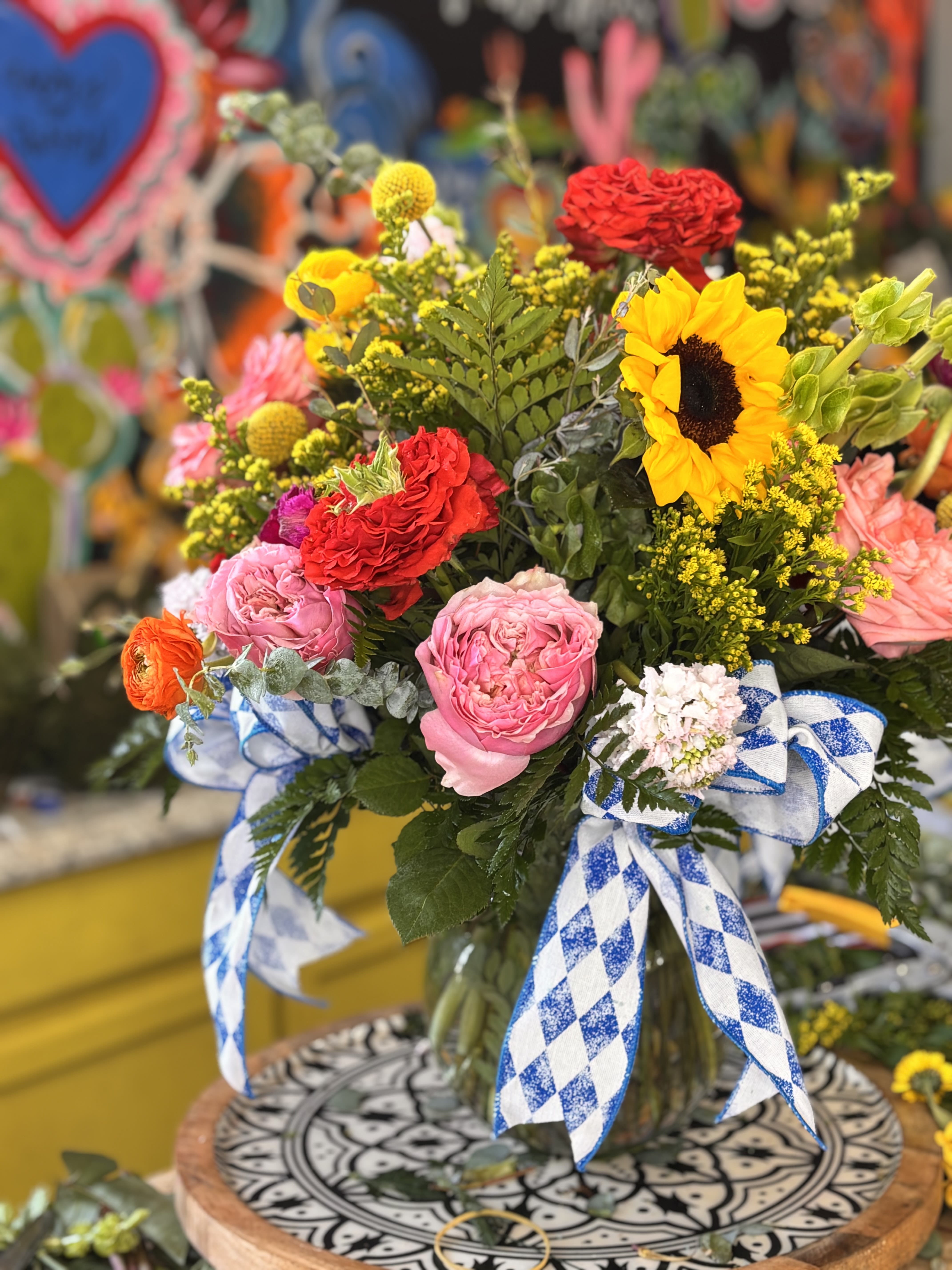 The Annie  - A bold, cheerful mixed-color flower arrangement that instantly reads as celebration - perfect as a birthday bouquet, congratulations gift, or a sunny just-because surprise. The Annie centers on a bright sunflower alongside pink roses and red carnations so the person you're thinking of will feel noticed and uplifted the moment it arrives.  Dense, layered textures of yellow filler blooms and varied greenery give this arrangement lively depth, while a blue-and-white ribbon bow lends a festive, folk-inspired accent. Finished in a clear glass vase and presented on a decorative tray, the bouquet balances warm reds and yellows with softer pinks for strong visual contrast and plenty of presence on a table or desk.  Send this when you want someone to feel celebrated, joyful, and genuinely seen - it's the kind of arrangement that announces the occasion without words. Vase included; arrange in a cool spot away from direct sun and replenish water daily to keep blooms looking fresh.