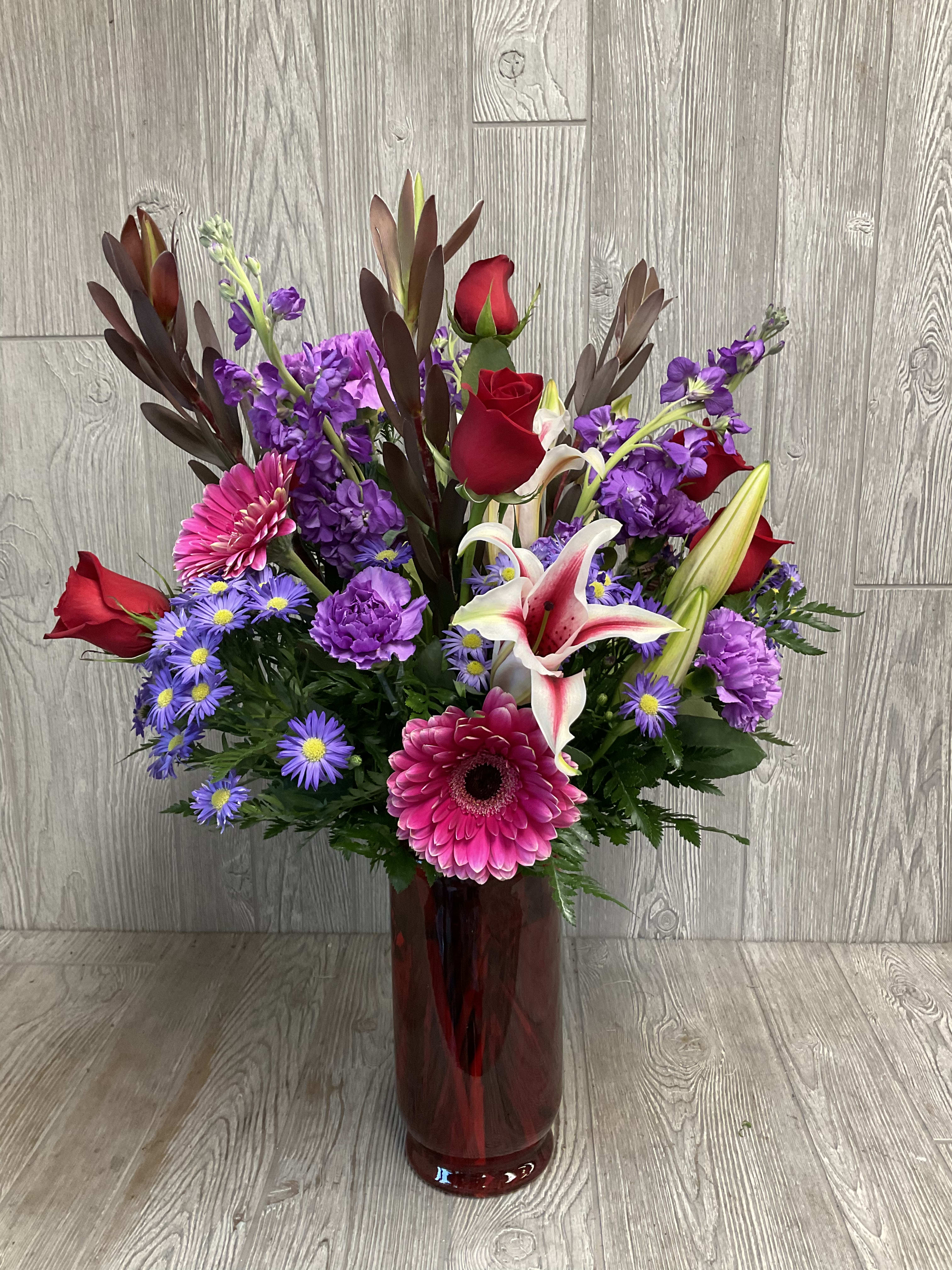 Mixed bouquet of red roses, lilies, and purple flowers in a red glass vase