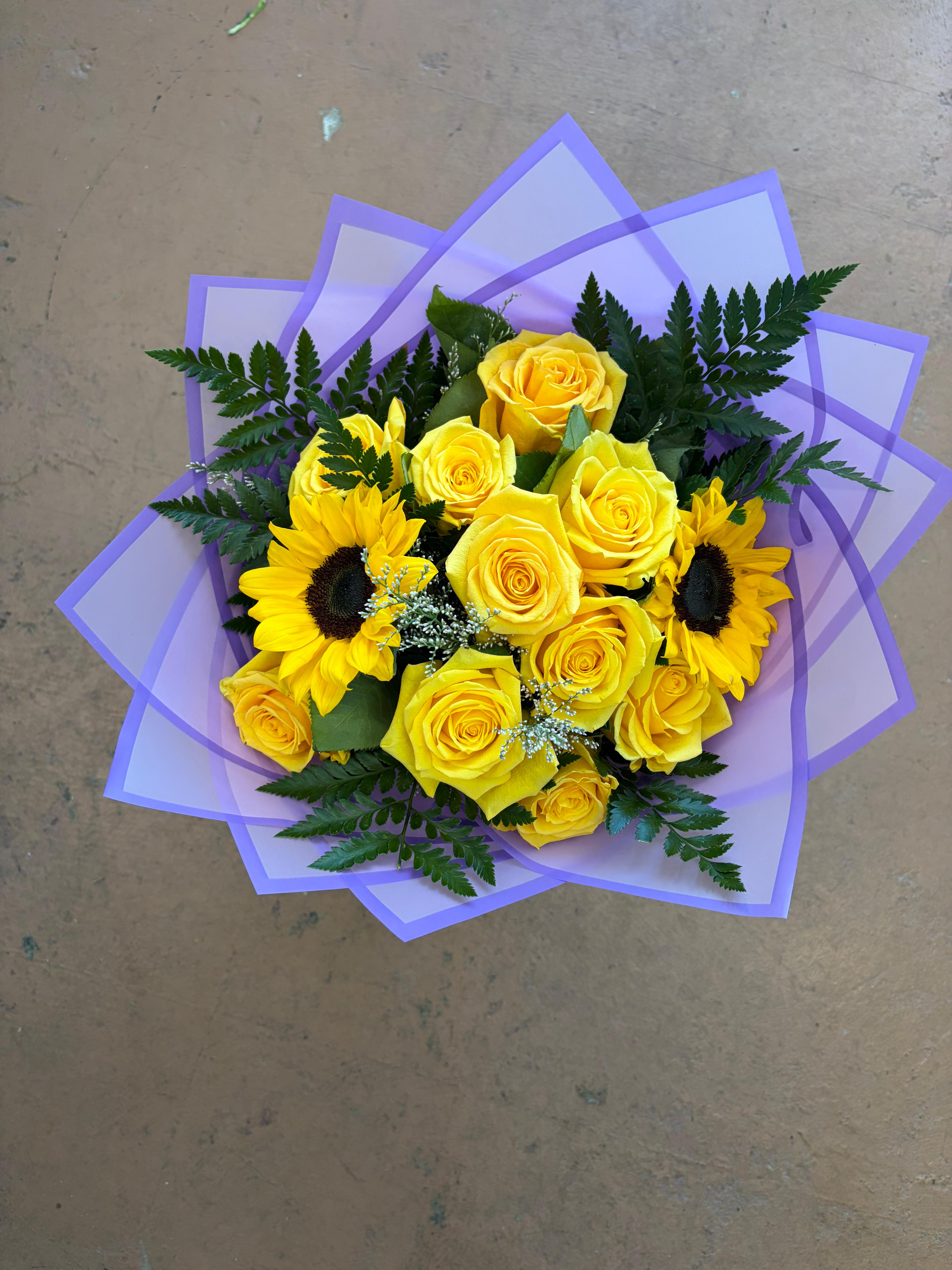 Golden Hour Bliss Bouquet - Bring the warmth of a summer afternoon into any space with our Golden Hour Bliss Bouquet. This radiant arrangement features a stunning combination of sun-drenched yellow roses and cheerful sunflowers, perfectly balanced to evoke feelings of joy and friendship. Hand-wrapped in a modern, layered lavender and purple wrap, this bouquet makes a bold statement that is sure to brighten someone's day.