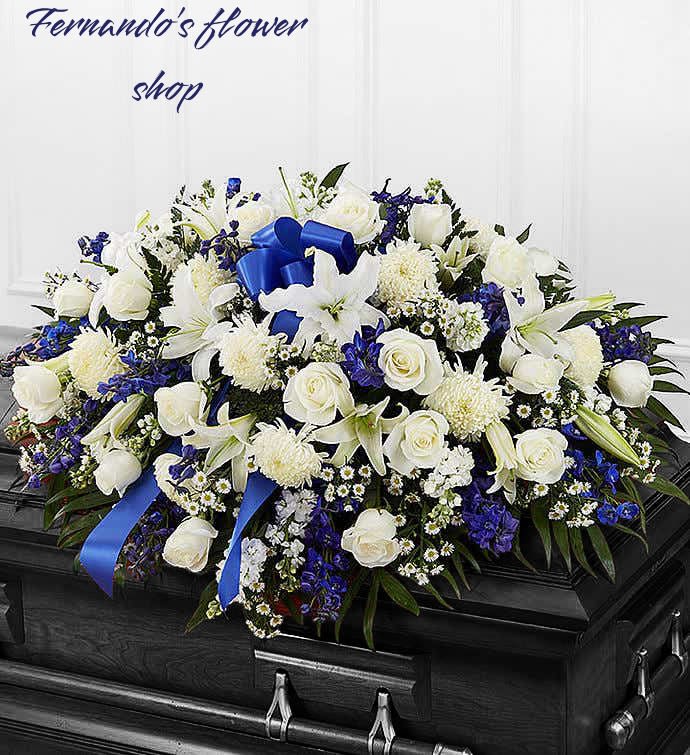 White and blue funeral casket spray with roses, lilies, and ribbon on a dark wood casket