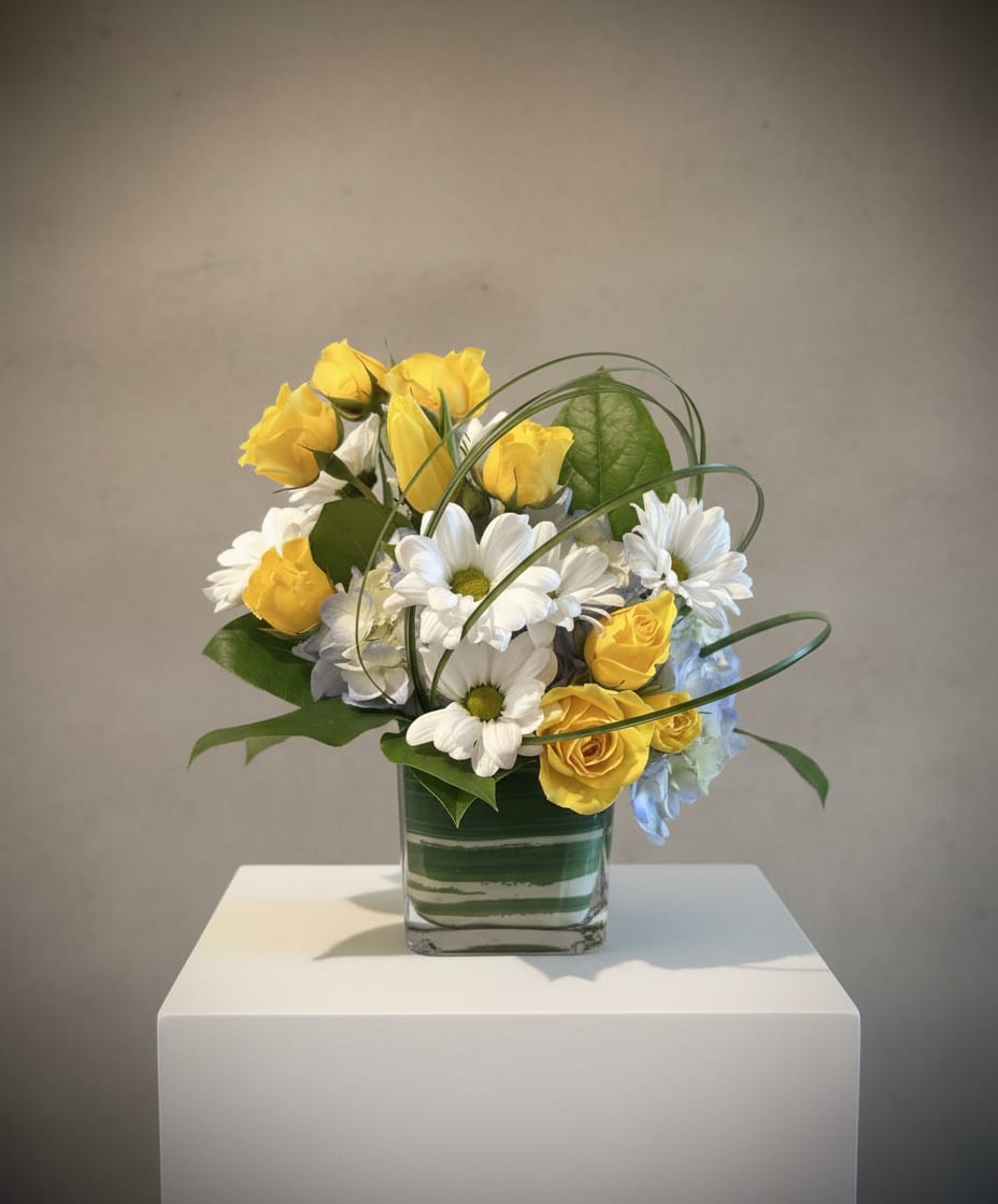 Thanks a Bunch! - Bright, cheerful yellow roses meet crisp white daisies and pale blue hydrangea accents in a compact glass vase-an uplifting flower arrangement perfect as a thank you bouquet, birthday surprise, or spring celebration centerpiece. The mix of sunny yellow and clean white balanced by soft blue creates an immediate sense of warmth and gratitude, the kind of gift that makes the person you're thinking of feel genuinely appreciated.  The design pairs classic roses, daisy blooms, and airy hydrangea clusters for textural contrast: full, velvety rose heads, delicate daisy centers, and cloud-like hydrangea petals. Long looping blades of decorative grass arc over the arrangement, adding light, airy movement that contrasts the denser blooms. Arranged low in a clear glass vase, it's compact enough for a desk, bedside table, or dining nook while still delivering a fresh, garden-picked look.  Ideal when you want to say "thank you" or brighten someone's day, this arrangement suits coworkers, friends, teachers, or hosts-anyone who should feel noticed. The glass vase makes for easy display; refresh the water every 2-3 days and trim stems slightly to keep blooms perky.