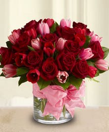 Round arrangement of red roses and pink tulips in a clear glass vase with a pink ribbon bow