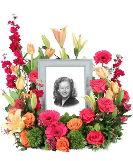 Consoling Embrace Memorial Flowers - Consoling Embrace is a stunning memorial arrangement brimming with hot pink roses and gerberas, complemented by peach tulips and lilies. Each bloom exudes radiant energy, symbolizing the enduring spirit of a loved one. As vibrant hues intertwine, this arrangement is a testament to the joy and beauty they have brought  Includes: Round Mache Wreath, Cut In Half With Halves Placed Next To Each Other, Italian Ruscus, Green Trick Dianthus, Hot Pink Gerberas, Green Hypericum, Peach Lilies, Hot Pink Roses, Purple Stock, Peach Tulips, Pink Waxflower, Orange Roses.