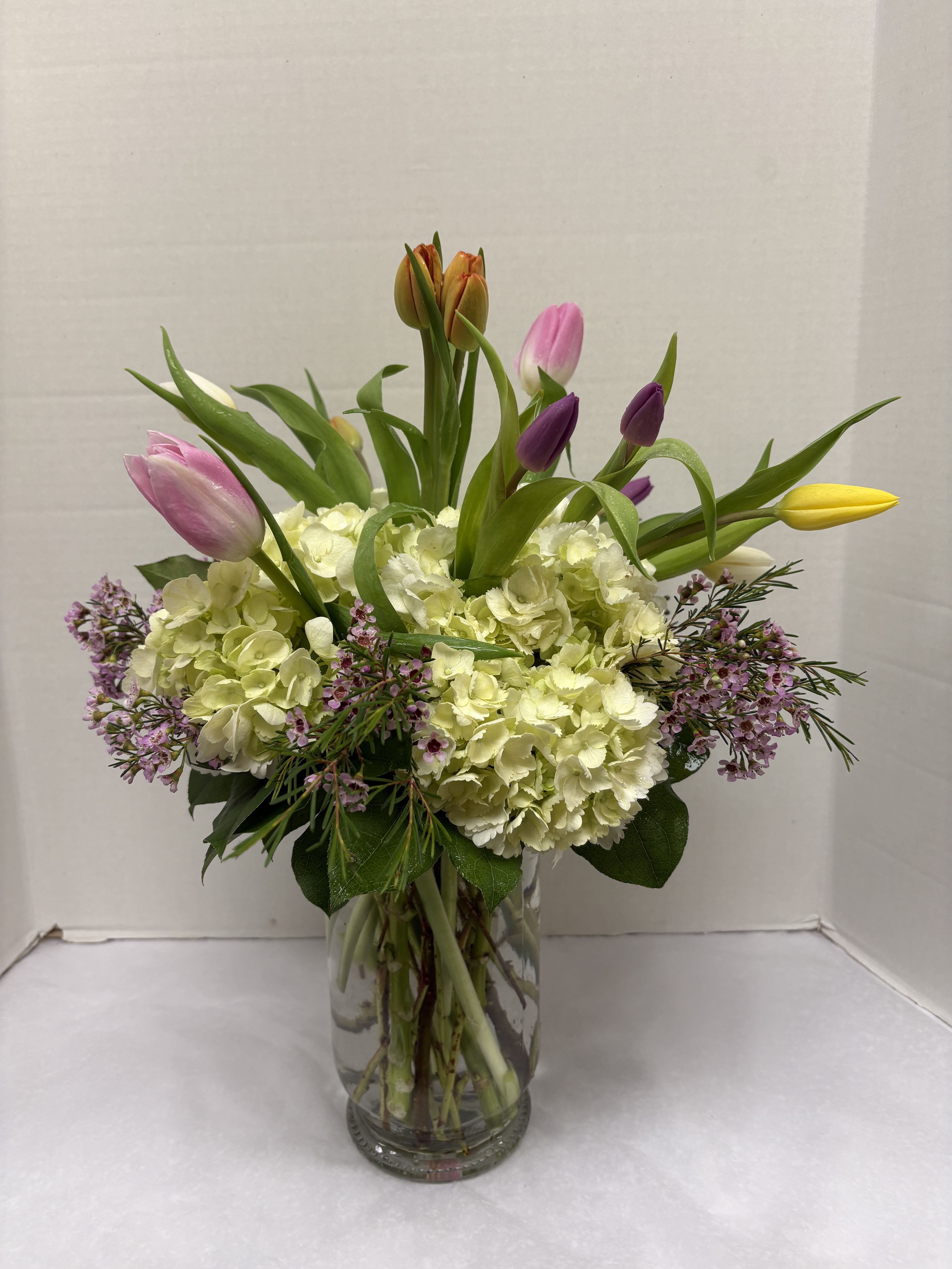 Timeless tulips  - A luminous cushion of pale ivory hydrangeas forms the foundation of this flower arrangement, while tall tulip stems in pink, purple, yellow, lavender and white rise above for a fresh, springlike silhouette - a perfect birthday bouquet, get well arrangement, or just-because surprise that reads as both elegant and joyful. The vertical tulips give motion and height so the person you're thinking of feels genuinely celebrated the moment it arrives.  Clear glass vase holds a dense rounded base of hydrangeas with delicate clusters of light pink and lavender filler blooms and fine-textured foliage softening the edges. Stems are gathered to show clean lines through the vase; colors include soft pastels and brighter yellow and purple accents for a cheerful mix.   Suitable year-round but especially lovely for spring and occasions like Mother's Day, Easter, anniversaries, housewarming, or Father's Day, this arrangement also works for sympathy, thank-you gifts, graduations, and weddings when a light, uplifting palette is wanted. Care tip: keep water fresh and trim stems every few days to extend vase life and keep blooms looking their best.