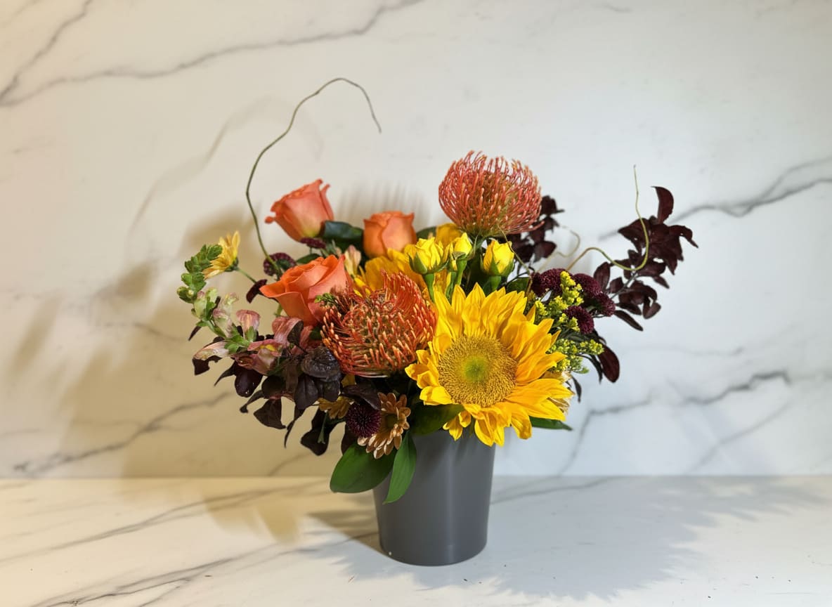 Bohemian Harvest - Celebrate the warmth and richness of the season with Bohemian Harvest, a free-spirited arrangement inspired by nature's autumn palette. Cheerful sunflowers take center stage alongside warm peach roses, striking pincushion protea, seasonal chrysanthemums, and textured accent blooms, all nestled among lush greenery and natural foliage. Designed with organic movement and earthy charm, this handcrafted bouquet captures the beauty of crisp fall days and abundant harvests.  Perfect for birthdays, housewarming gifts, Thanksgiving gatherings, or simply bringing the cozy colors of autumn into your home, Bohemian Harvest is a vibrant expression of the season's natural beauty.