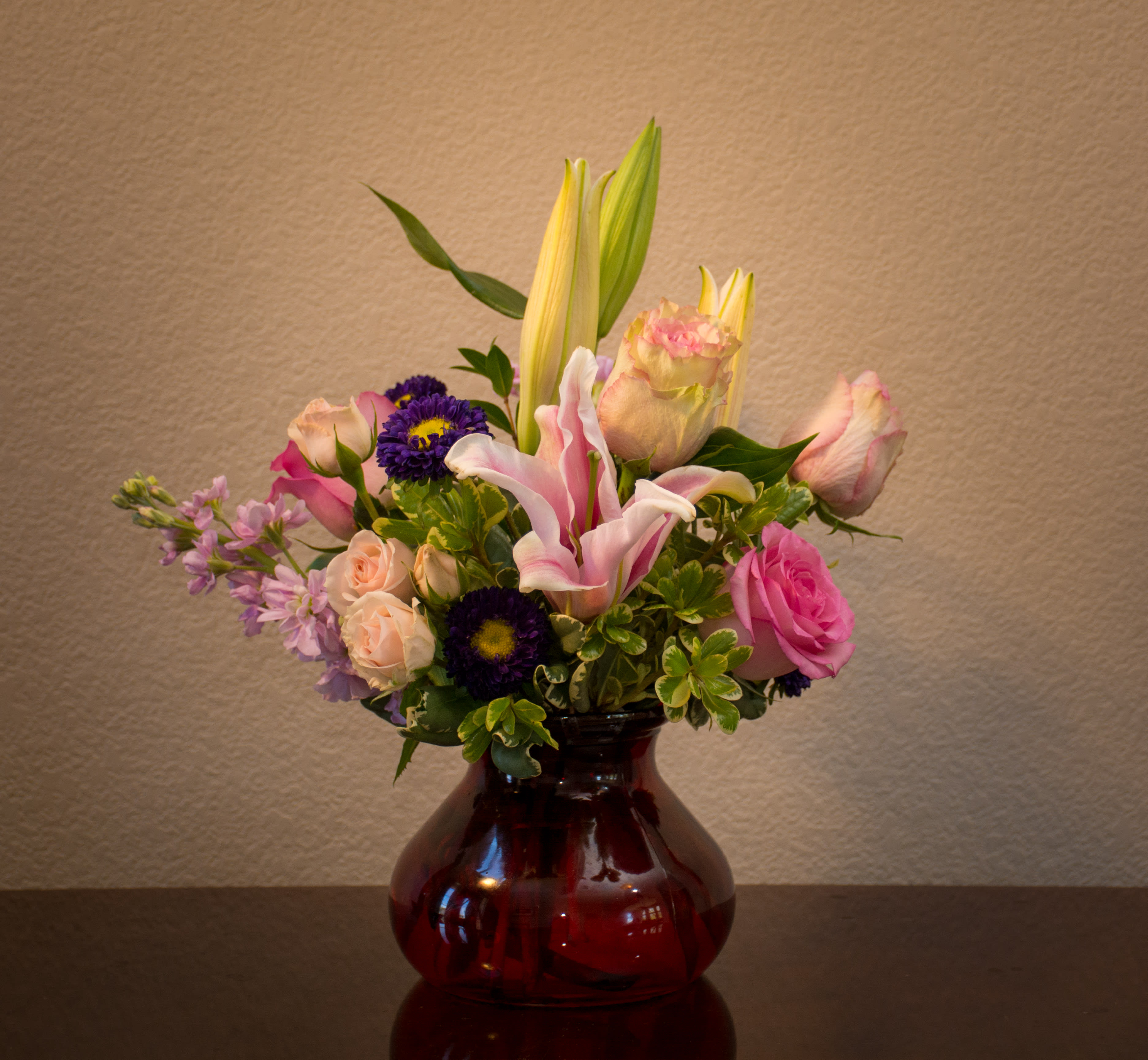 Mixed bouquet of lilies, roses, and purple daisies in a red glass vase