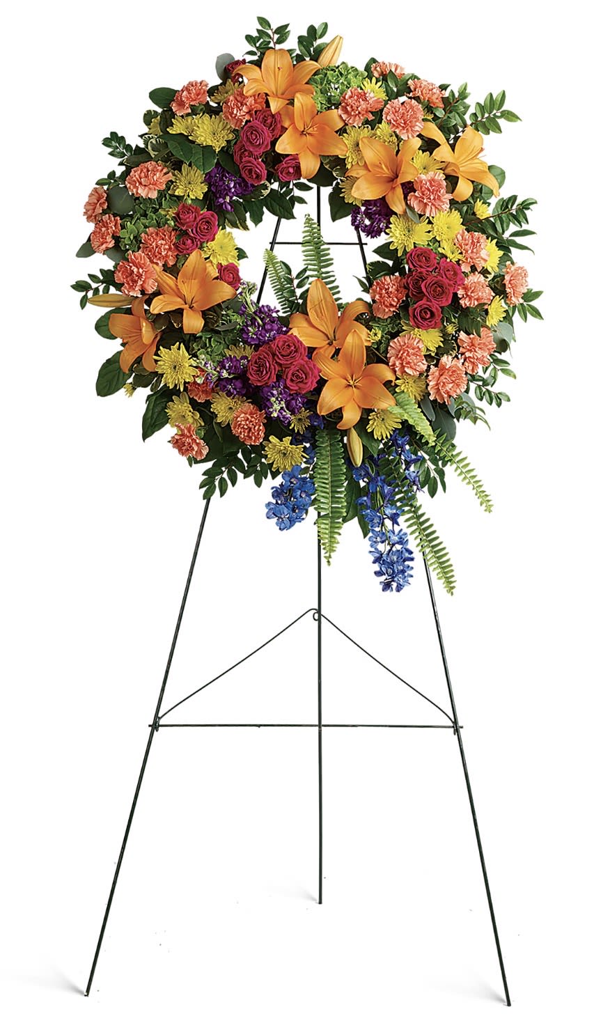 Colorful Serenity Wreath - A vibrant circular wreath that brings a sense of comforting brightness to a sympathy or funeral setting, this large standing wreath pairs bold orange lilies with soft pink roses to create an immediate, uplifting impression. The product type and occasion - a funeral sympathy flower arrangement on an easel - is clear from the start: the elevated, formal presentation gives the piece ceremonial weight while the saturated mixed-color palette reads as both respectful and hopeful for those receiving it.  Crafted with lilies, roses, carnations, chrysanthemums, delphinium and other seasonal blooms, the wreath mixes coral carnations and sunny yellow chrysanthemums with purple accents and trailing blue flowers for depth and movement around the ring. The vivid combination of orange, pink, yellow, blue, red and peach tones creates textural contrast between the large lily blooms and the clustered roses and chrysanthemums; the wreath is mounted on a sturdy black metal easel so it stands prominently at view.  Made to honor and to celebrate a life, this arrangement is the kind of tribute that helps the recipient feel seen and remembered - respectful for a memorial service yet bright enough to reflect a joyful personality. Please note the standing design arrives ready to display on its metal easel; for longevity, keep flowers in a cool, sheltered area away from direct sun and heat so the colors and form hold through services.