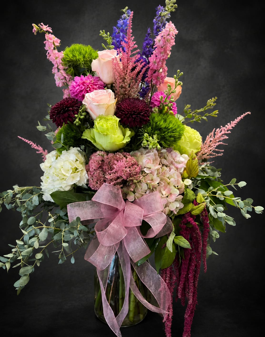 Sweet Overflow - Pastel pink roses and cushiony hydrangeas meet vivid purple delphinium spikes in this tall, layered flower arrangement-a birthday bouquet that reads as both romantic and joyously celebratory. Sweet Overflow's vertical silhouette and trailing greenery create a full, dramatic presence that makes the person you're thinking of feel truly seen-perfect for birthdays, anniversaries, or a spring surprise that says "you matter."  Built in a clear glass vase and finished with a sheer pink ribbon, the design mixes rounded blooms (roses, hydrangeas, chrysanthemums) with upright stock and delphinium accents for texture and movement. Bright chartreuse-green florals and deep burgundy chrysanthemum-like flowers add contrast against airy eucalyptus-style foliage, producing a lush, polished composition that works equally well for get well, housewarming, thank you, thinking of you, I'm sorry, or just because moments.  This arrangement arrives gift-ready in a glass vase with a large pink bow-no extra wrapping needed-making it an easy, impressive choice for love and romance, congratulations, Boss's Day, or Admin Professionals' Week. Size and fullness give it strong presence on an entry table or bedside, and simple care (fresh water, a cool spot out of direct sun) will keep the blooms looking their best through the celebration.