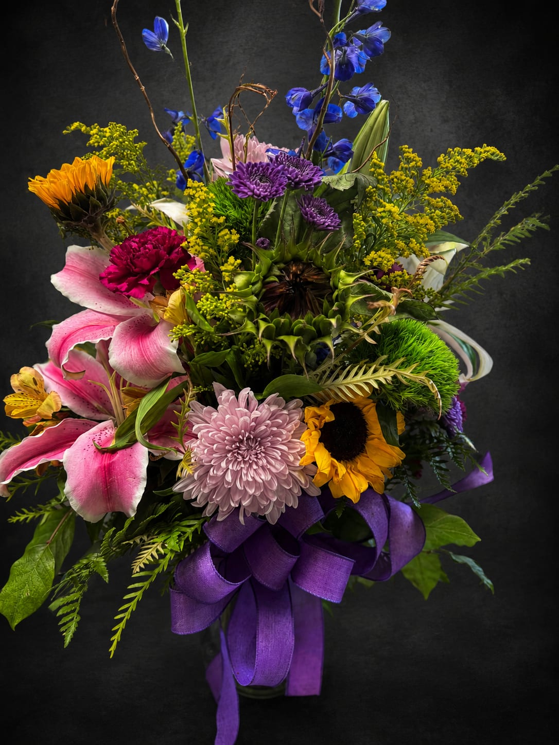 Sun Delight - A tall, garden-style mixed bouquet that greets the eye with cheerful sunflowers and fragrant pink lilies-perfect as an anniversary bouquet or a bright "thinking of you" surprise. Sun Delight reads as celebratory and warm: the bold yellow sunflowers and pops of purple make an immediate impression, while the layered composition and airy stems give the arrangement the kind of presence that makes someone feel truly noticed.  This large arrangement combines pink lilies, yellow sunflowers, purple chrysanthemums and daisy-like blooms, blue accents, and clusters of small yellow filler flowers for a textured, abundant look. It arrives tied with a wide purple ribbon and presented in a vase, so the recipient can enjoy the display right away. The mix of warm and cool tones-pink, purple, yellow and green-keeps it fresh for summer but just as fitting for get well wishes, housewarming, thank yous, or a cheerful "just because" moment.  Designed to lift spirits and celebrate milestones, Sun Delight makes a thoughtful gift for someone recovering at home, settling into a new place, or marking a special anniversary or work appreciation moment like Boss's Day or Admin Professionals' Week. Vase-arranged and ready to display; trim stems and refresh water as needed to extend bloom life.