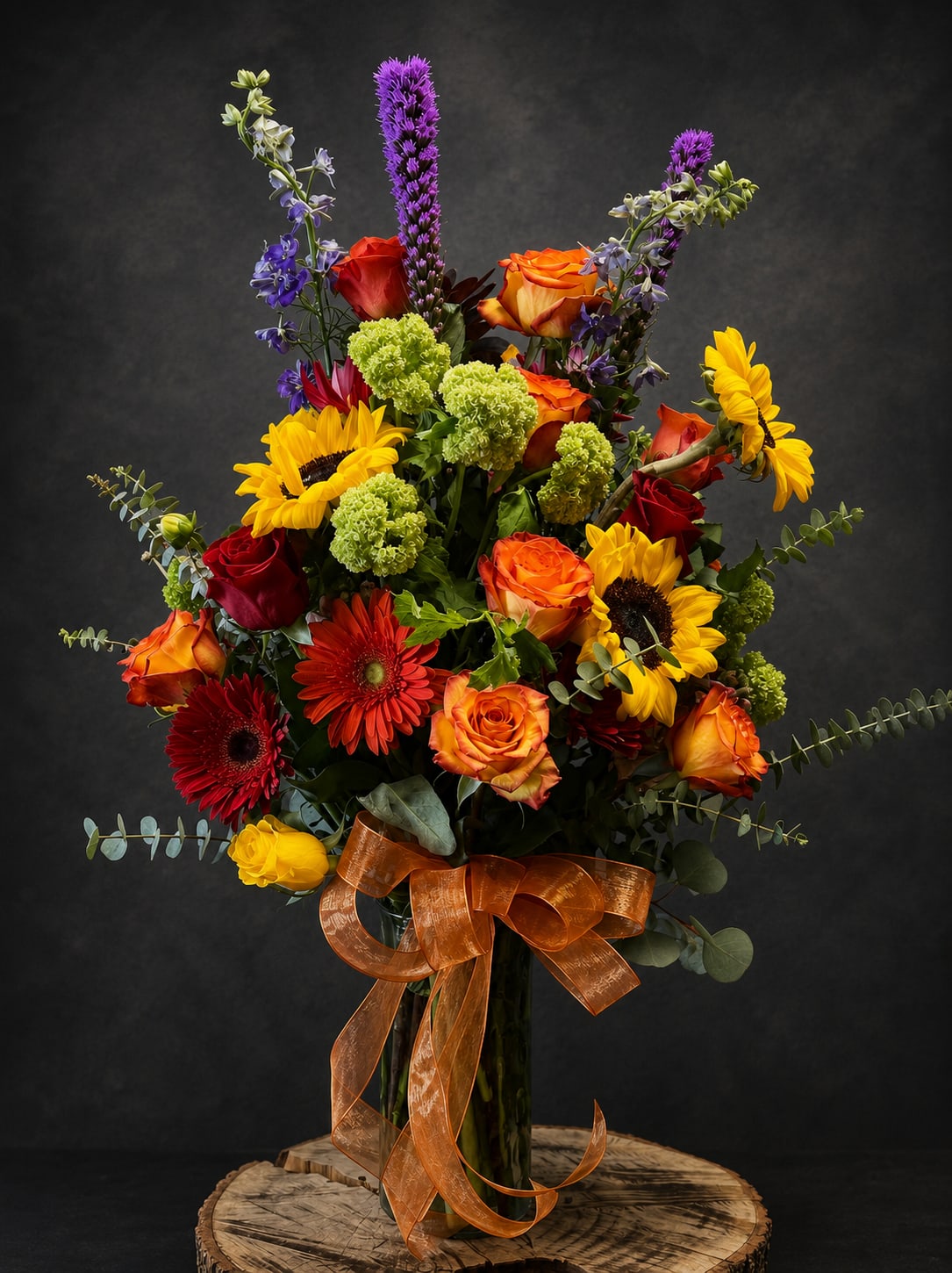 Forever Surprised  - Bold, sunlit color greets the recipient with this tall mixed flower arrangement-perfect as an anniversary bouquet, birthday surprise, or a cheerful "thinking of you" gift. The combination of orange and red roses, bright yellow sunflowers, and red gerbera daisies creates an immediate warm, celebratory impression, while purple delphinium spikes and lime-green pom‑pom chrysanthemums add verticality and playful contrast that make the arrangement feel both abundant and refined.  Arranged in a clear glass vase and finished with a wide translucent orange ribbon tied around a wooden base, the design reads as polished yet joyful. Textures range from the smooth rose petals to the bold sunflower faces and the airy eucalyptus and vertical delphinium, giving the bouquet movement and depth. This tall composition works well for housewarming, congratulations, retirement, or to lift spirits for get-well and sympathy moments without feeling overbearing.  Whether you're sending thanks, saying "I'm sorry," celebrating Mother's Day or Boss's Day, or simply brightening an autumn or winter day, this arrangement sends a warm, heartfelt message that the person you're thinking of will feel genuinely seen. The clear vase presentation arrives ready to display; for best longevity, refresh the water and trim stems every few days.