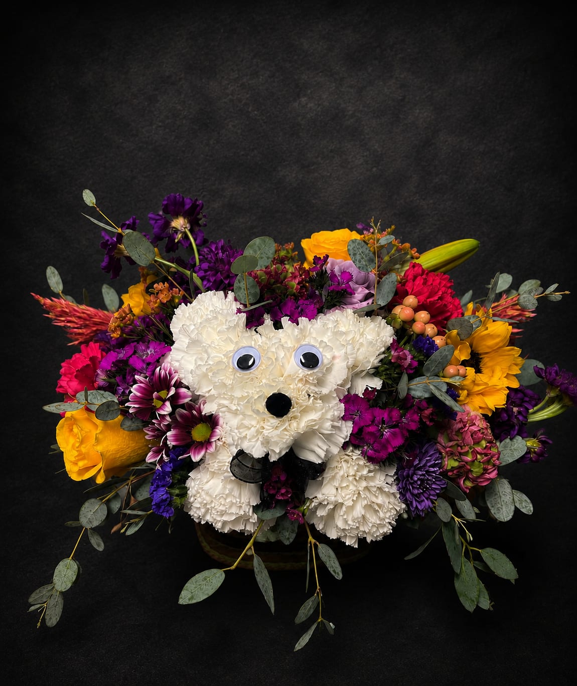 Buzz Dog Bouquet - A playful white teddy-bear shaped flower arrangement that reads like a joyful surprise-perfect as a birthday bouquet, a thank-you gift, or a thinking-of-you pick-me-up. The soft white body formed from densely packed carnations creates an unmistakable cuddly silhouette, while small black accents and a black nose decoration give the bear a sweet, expressive face that will make the recipient smile.  Surrounding the carnation bear is a bright, rounded halo of mixed flowers in purple, yellow, pink and orange-roses, chrysanthemums, daisies and other mixed blooms blend with ruffled textures for lively contrast. Googly eyes and a basket presentation add novelty charm, and trailing eucalyptus brings movement and a touch of green to the dense composition. The overall size and fullness read as a giftable centerpiece that looks great on a table, desk, or entryway.  Whether you're saying happy birthday, sending thanks, or simply letting someone know you're thinking of them, this whimsical animal arrangement delivers cheerful personality and warmth. It's an ideal choice for children, dog lovers, or anyone who appreciates a lighthearted floral surprise-order for same-day delivery when available to make their day instantly brighter.