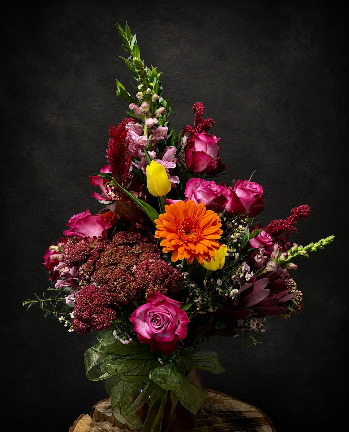 You and Me - A dramatic, upward-sweeping bouquet that reads like a celebration in color-vivid pink roses, a single bright orange gerbera, and sunny yellow tulips create an instant focal point while tall snapdragon-like spikes lift the arrangement with elegant vertical movement. This flower arrangement for anniversaries, birthdays, get well wishes, or romantic gestures makes the person you're thinking of feel noticed and celebrated the moment it arrives.  Layered and lush, the design mixes saturated pinks, warm reds, oranges and yellow with burgundy and pale pink accents for high-contrast, energetic appeal. The stems are arranged in a clear glass vase and finished with a sheer green ribbon for a polished, hand-tied look; the composition reads tall and formal but still warmly accessible. Ideal for housewarming, thank-you, "just because," I'm sorry, Mother's Day or Thinking of You moments, it also suits workplace occasions like Boss's Day or Admin Professionals' Week-anytime you want someone to feel truly seen.
