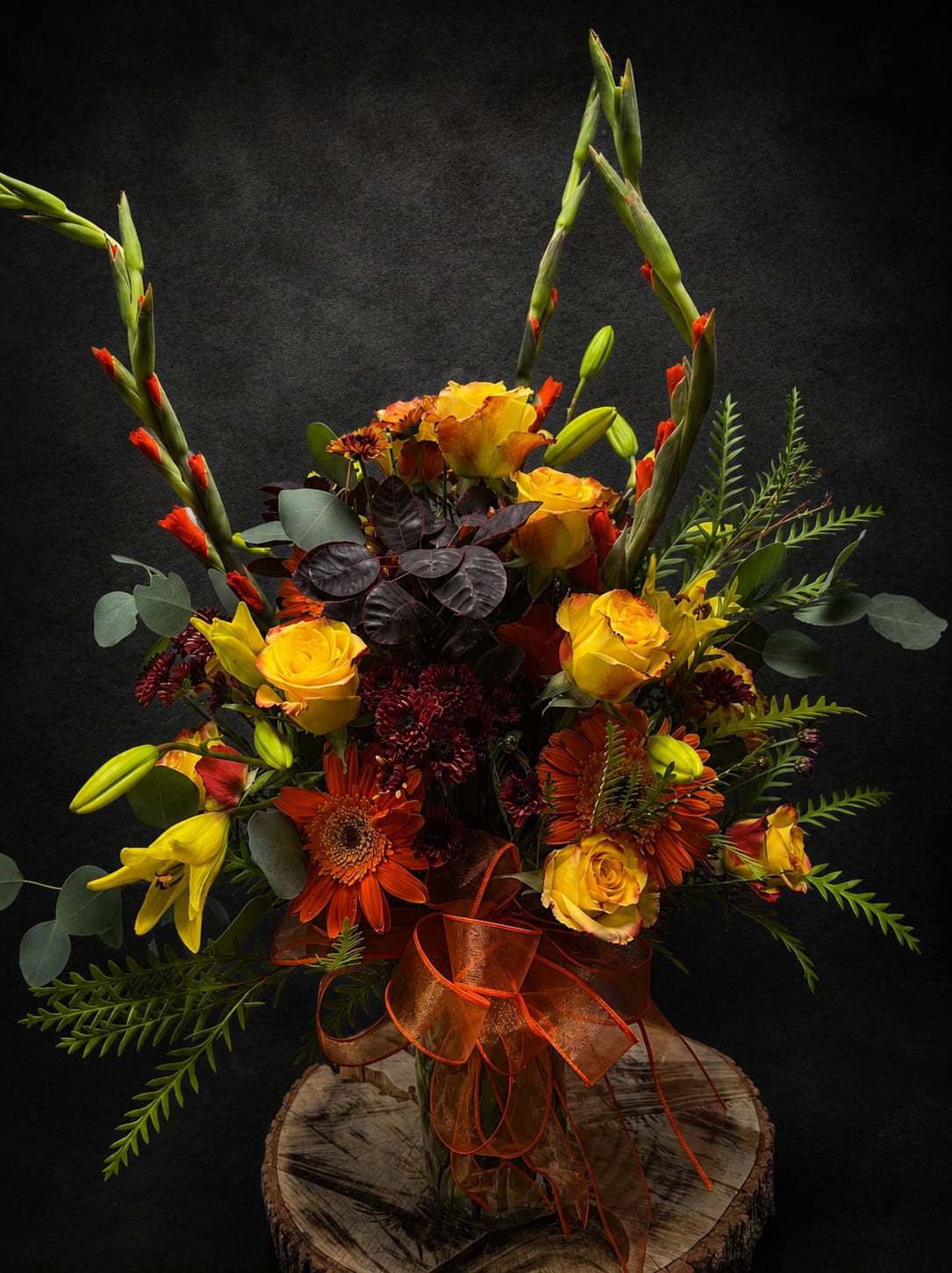 Autumn Sunrise - A tall, sculptural autumn bouquet that reads like a sunrise-rich yellow roses, bright orange gerbera daisies and bold red chrysanthemums gather in a dense central cluster while long gladiolus-like spikes soar above. This flower arrangement is a flower arrangement for anniversary, birthday, Thanksgiving or "just because" moments when you want the person you're thinking of to feel celebrated and seen; its warm red, orange and gold palette also makes a striking sympathy or thank-you tribute with a formal, respectful presence.  The design combines sunflowers, lilies, stock and mixed seasonal blooms with airy greenery and dark burgundy foliage for dramatic contrast and texture. It arrives in a clear glass vase set on a wood slice base, finished with a sheer orange ribbon for an autumnal flourish. The vertical silhouette and dense center give this piece a sculptural, gallery-like quality that reads well on a mantel, entry table, or beside a bedside at a funeral service.  Perfect for autumn occasions-Thanksgiving gatherings, anniversary dinners, or as a bold birthday surprise-this arrangement also translates to Grandparent's Day, Father's Day, Women's Day, Boss's Day or congratulations gifts. Care is simple: keep water fresh, trim stems every few days, and place away from direct heat to extend blooms; choose delivery or pick-up at checkout to ensure it arrives at the right moment.
