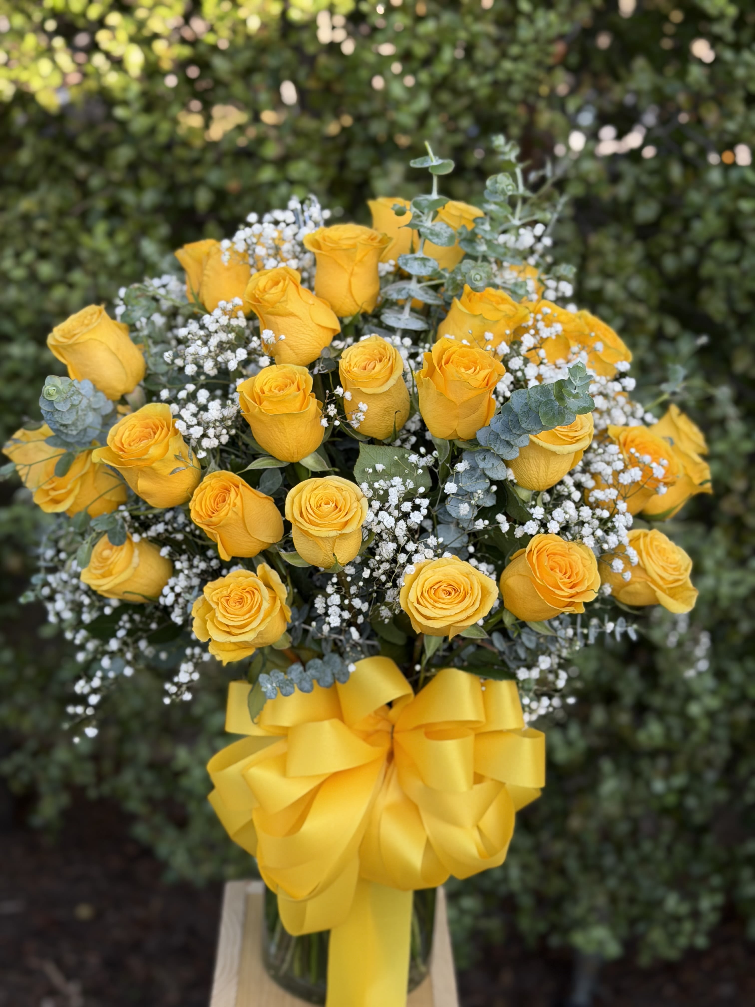 Three Dozen Yellow Roses  - A dense cloud of three dozen bright yellow roses creates an immediate sunny impression-perfect as a birthday bouquet or a cheerful "just because" gift that reads romantic as well. The strong yellow palette and layered yellow ribbon make her feel celebrated and seen, whether you're marking spring milestones, saying congratulations, or sending a warm note of love and romance.  Each arrangement pairs the vivid roses with small white filler blooms and silvery-bluish eucalyptus for contrast and texture, arranged low and full in a clear glass vase and finished with a large yellow ribbon. The piece arrives on a simple wooden stand for a polished presentation; the compact, lush shape and durable glass vessel make it easy to display. Ideal for Mother's Day, an anniversary, or an everyday pick-me-up-refresh water every other day and trim stems as needed to keep the bouquet looking its brightest.