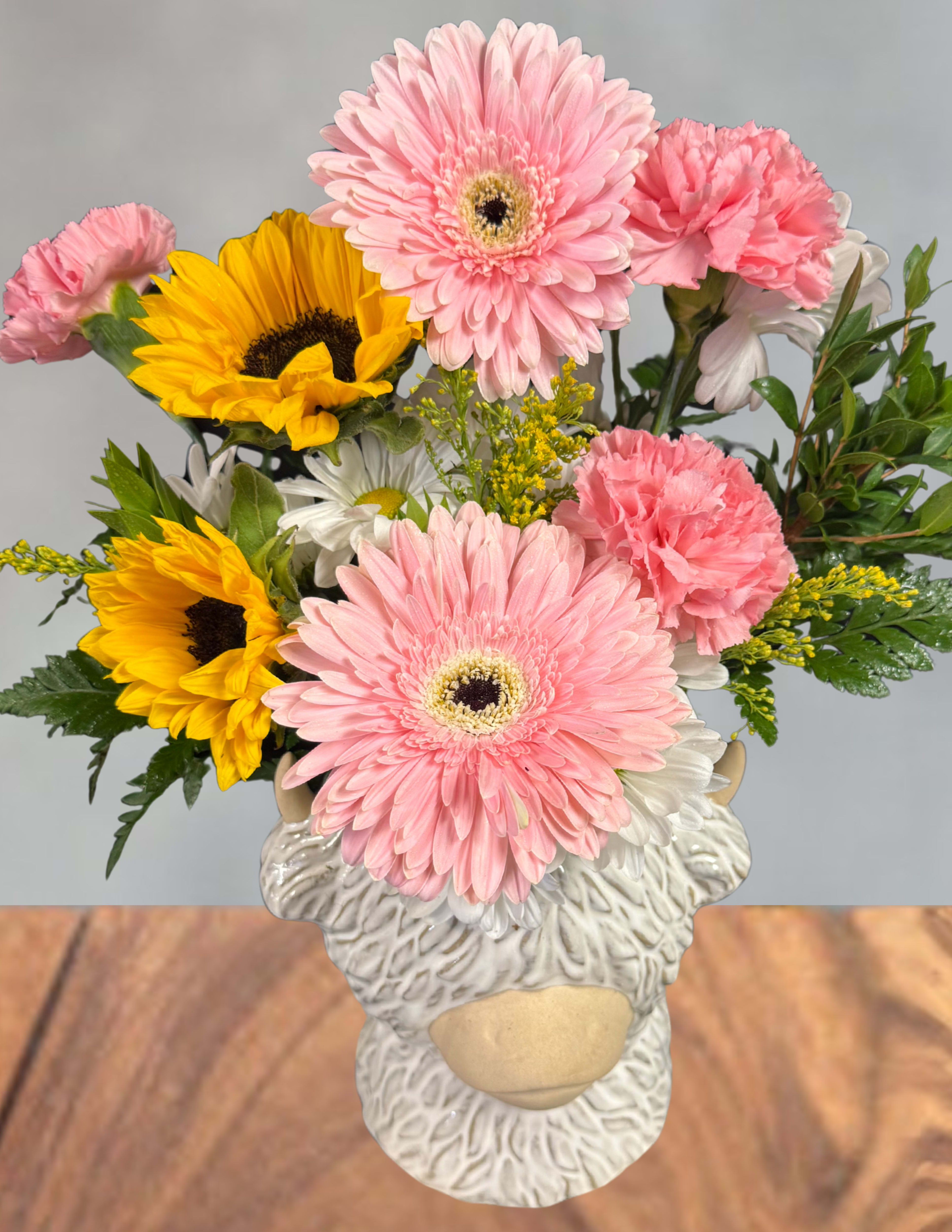 Highland cow flower arrangement  - Bright, playful and impossible to miss, this Highland cow flower arrangement is a birthday, thank-you, or just-because bouquet that will make the person you're thinking of smile. The whimsical ceramic sheep/cow planter-textured, hand-finished in soft white and tan with little horns and a beige snout-gives the gift a rustic, novelty charm while the loose, casual composition reads as joyful rather than formal.  Inside the planter, large pink gerbera daisies mingle with vivid yellow sunflowers, soft pink carnations, and crisp white daisies, finished with touches of yellow filler and fresh green accents. The contrast between soft pinks and bright yellows creates a cheerful color story that feels lively and friendly; the flowers are loosely clustered for a natural, hand-picked look. The arrangement arrives in the ceramic sheep/cow vase shown, ready to display on a table or windowsill.  Perfect for brightening a kitchen, office, or bedside table, this arrangement is ideal for someone who enjoys a bit of whimsy with their blooms-whether celebrating a birthday, saying thanks, or sending a spontaneous pick-me-up. Care is simple: keep flowers in clean water, trim stems every few days, and place away from direct heat to extend their lively, fresh appearance.