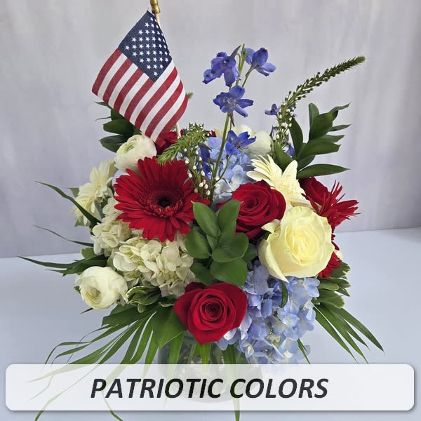 Patriotic red, white, and blue flower arrangement with roses, gerbera daisies, hydrangeas, delphinium, and a small American flag.