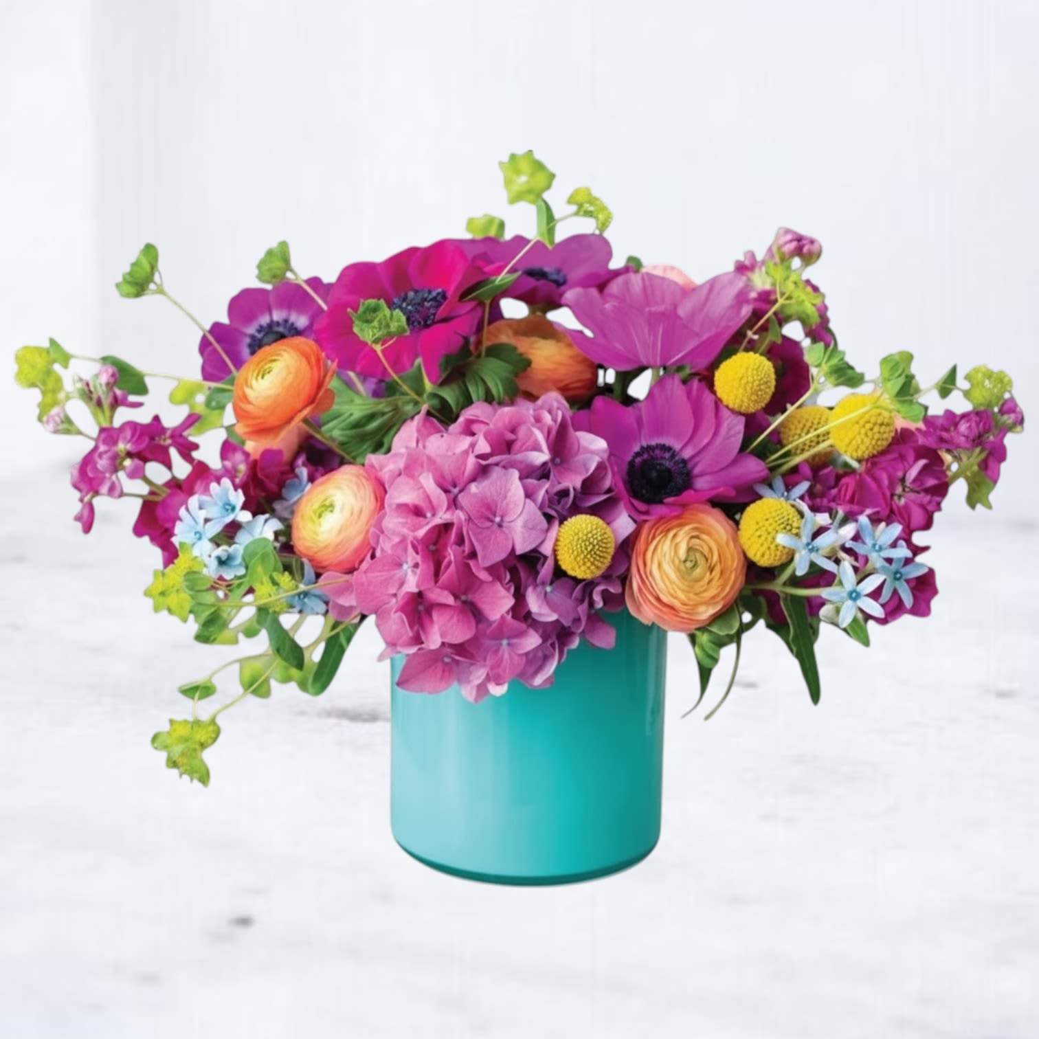 Picturesque - Vivid pink roses and ranunculus take center stage in this birthday flower arrangement, surrounded by peach and yellow ranunculus for warm contrast-perfect for a birthday bouquet or a bright table accent. A cloud-like head of purple hydrangea anchors one side while blue and orange notes lift the palette; the overall effect reads joyful and modern, made to make the person you're thinking of feel celebrated and truly seen.  This arrangement combines hydrangeas, ranunculus, and roses in shades of pink, purple, yellow, orange, and blue, accented with small green filler flowers and rounded foliage for a lush, layered look. Blooms are arranged compactly in a smooth turquoise ceramic cube vase that adds a playful, contemporary touch and keeps the design feeling fresh on mantels, desks, or dining tables.  Ideal for birthdays, congratulations, special gatherings, or as a cheerful everyday gift, Picturesque arrives ready to enjoy; the compact shape works well for same-day delivery and for brightening small spaces. Gentle daily refreshing of the water and trimming stems every few days will help extend the life of the bouquet.