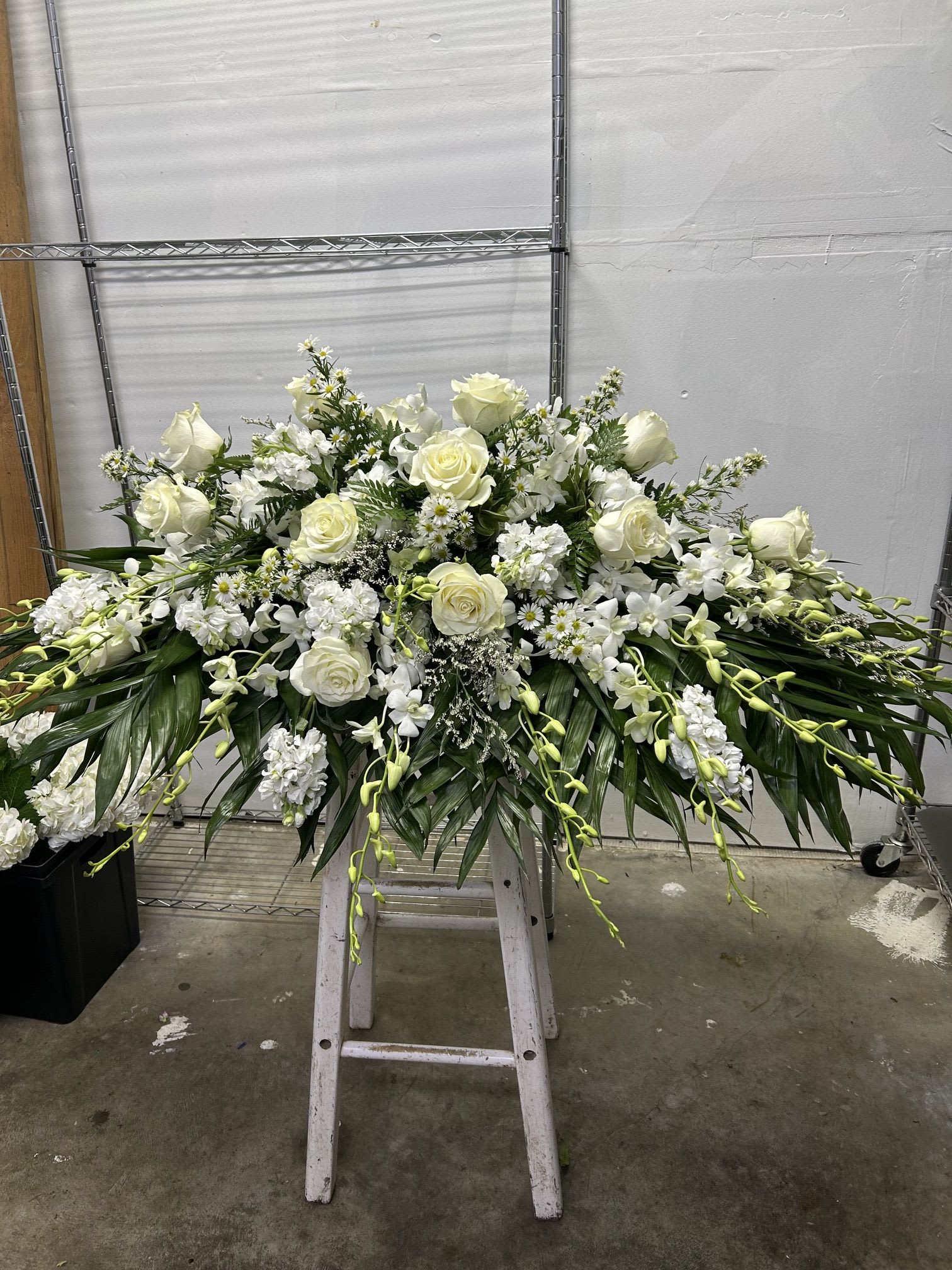 Peaceful Remembrance - A sweeping all-white funeral spray designed to offer calm and dignity, this Peaceful Remembrance arrangement is a formal memorial piece meant for a service or graveside tribute. The low, wide silhouette and the arrangement's placement on a floral stand give it the respectful scale and presence that help convey sympathy and honor at a funeral or memorial service.  Clustered white roses form the heart of the spray, surrounded by elegant white orchid sprays and small daisy-like filler blooms that add delicate texture. Long, arching greenery and trailing orchid stems extend outward from the center, creating a gentle, cascading profile that reads both full and refined. The neutral white-and-green palette keeps the mood solemn and peaceful while the mixed blooms provide visual depth and soft contrasts of shape and form.  Choose this piece when you want the person you're remembering to be honored with a thoughtful, formal presentation; it's the kind of tribute that helps mourners feel seen and supported. The arrangement arrives mounted on a floral stand and is sized for ceremony display-no special care is needed beyond keeping it sheltered from extreme heat or wind until the service.