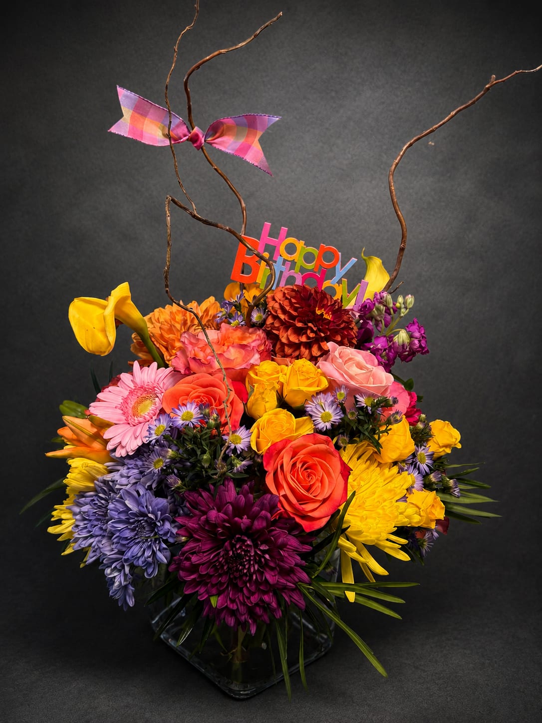 Birthday Surprise - A burst of party-ready color-this birthday bouquet is designed to make the person you're celebrating feel seen and special. As a birthday flower arrangement, it announces joy the moment it arrives: bright roses, vivid gerbera daisies, cheerful chrysanthemums and small daisy-like accents mingle with yellow calla lilies and pops of coral, pink, purple and red for a playful, festive look. A colorful "Happy Birthday" sign and bright plaid ribbon bow complete the celebratory presentation.  Built tightly in a clear glass vase, the design layers large focal blooms with smaller filler flowers for a full, lush silhouette while tall curly branches and twigs add height and whimsical movement. Decorative ribbons, a birthday pick and curly branches give it extra personality-perfect for someone who loves exuberant color and a lively display.  Ideal for friends, family members, coworkers, or anyone marking another happy year, this arrangement sends a joyful, celebratory message that's both thoughtful and fun. Comes ready to display in its clear vase; refresh water and trim stems as needed to keep blooms looking fresh for several days.