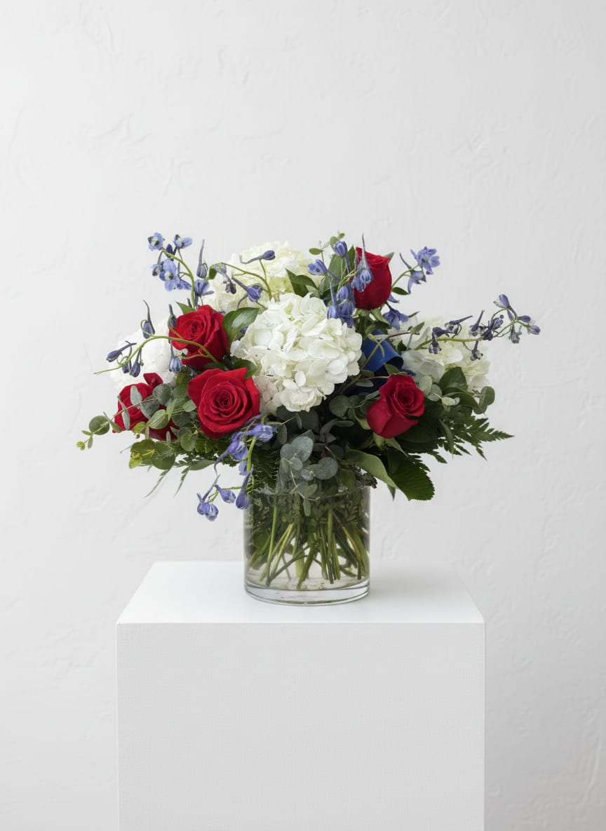 Patriotic Blooms                                           - Deep red roses paired with a full cluster of white hydrangeas and airy blue accents create a striking patriotic flower arrangement perfect for an anniversary, Valentine's Day, or any moment you want the recipient to feel honored and remembered. This flower arrangement brings a classic, romantic feel with a fresh, celebratory mood-the kind of bouquet that makes someone feel truly seen whether you're marking love, gratitude, or a respectful tribute.  Tightly gathered in a clear cylindrical glass vase, the composition reads full and rounded: deep red roses anchor the piece, crisp white hydrangeas add volume and softness, and delicate blue blooms lend lightness and contrast. Greens provide subtle texture and support, keeping the palette clean and balanced. The clear glass vase showcases the stems for a timeless look that fits mantelpieces, dining tables, or a bedside stand.  Ideal for gifting to a partner on an anniversary, sending warm patriotic sympathy, or celebrating a special Valentine, this arrangement arrives ready to display and impress. For best longevity, place in fresh water out of direct sunlight and change the water every few days to keep blooms looking their best.