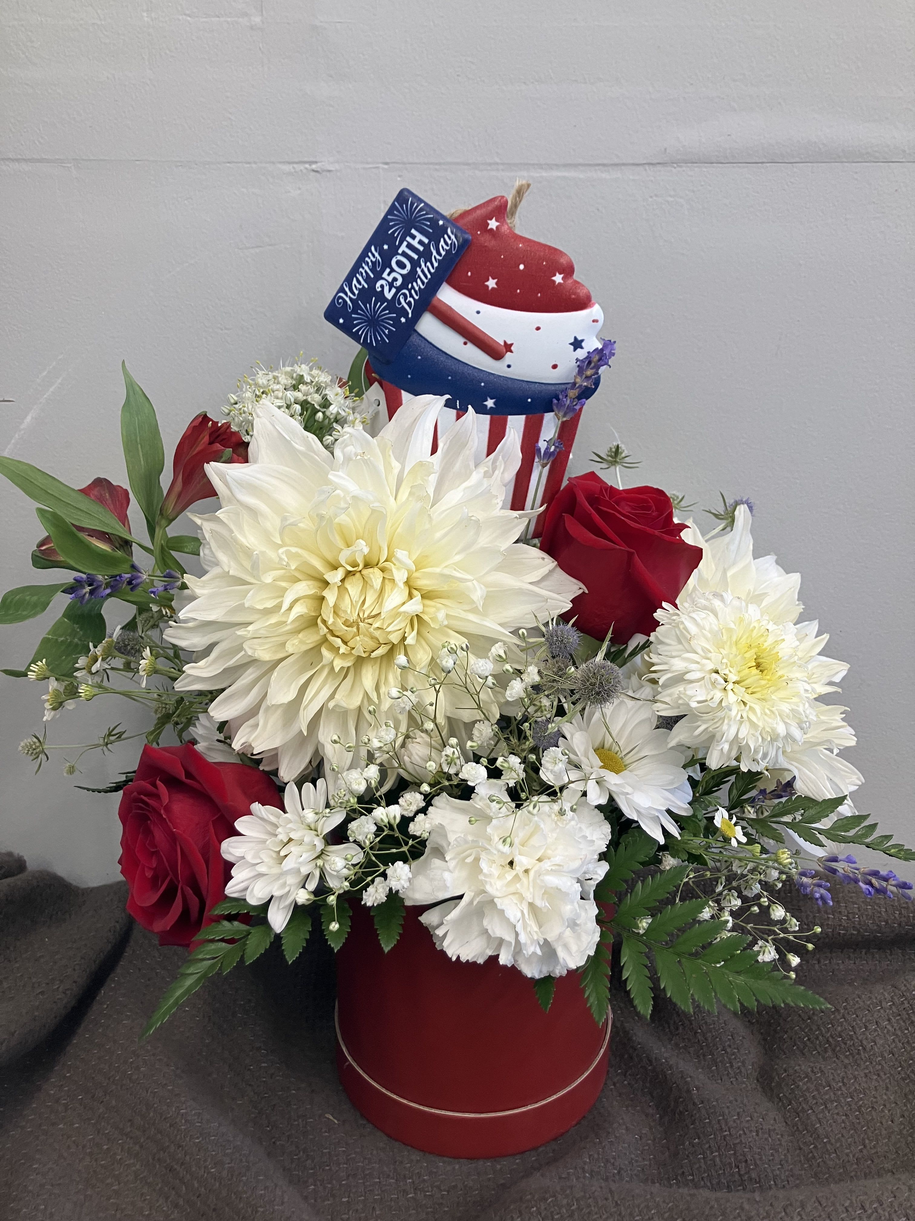 The Red White and Blue - Celebrate America’s 250 birthday with large white dahlias, red roses and blue thistle in a red barrel arrangement with a cute tin cupcake sign. 