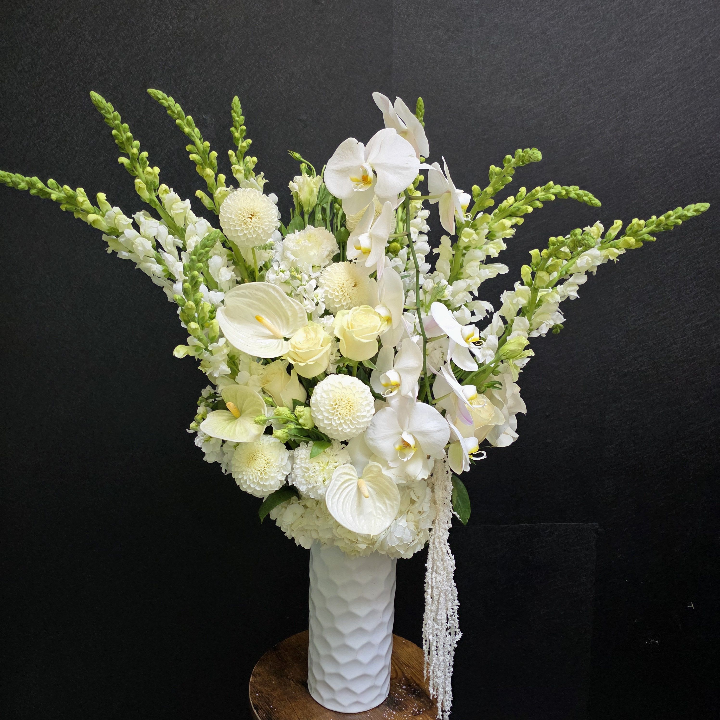 Moonlit Grace - Tall, sculptural white blooms make a serene first impression in this flower arrangement-ideal as a wedding centerpiece, sympathy tribute, or refined gift to mark a quiet, meaningful moment. The composition's strong vertical lines and arching orchid sprays give it an elegant, gallery-like presence that will make the person you're thinking of feel truly seen and honored.  Phalaenopsis orchids and calla lily forms dominate the palette, balanced by round chrysanthemum heads, delicate roses, and airy snapdragon spikes in bright green for height and contrast. A few anthurium-style blooms add smooth, modern texture, all arranged in a white textured ceramic vase that emphasizes the pale palette and clean silhouette. The arrangement reads formal yet fresh, with graceful movement from the orchid sprays and linear snapdragons.  Perfect for weddings, sympathy, or any moment when a polished, calming statement is called for-this design delivers quiet beauty and lasting impact. The tall, airy build suits display on pedestals, entry tables, or beside a mantle; no special care beyond fresh water and a cool location will help preserve the blooms' elegant form.