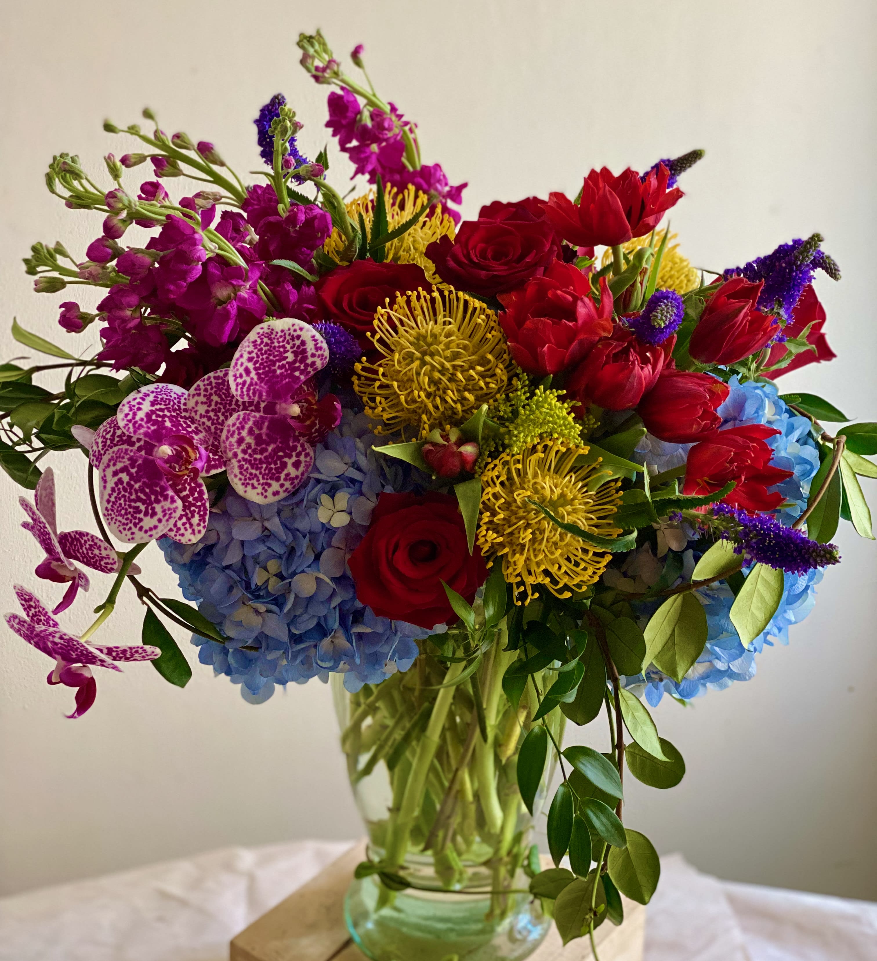 Tall mixed arrangement of red roses, yellow pincushion protea, blue hydrangeas, and spotted orchids in a clear glass vase