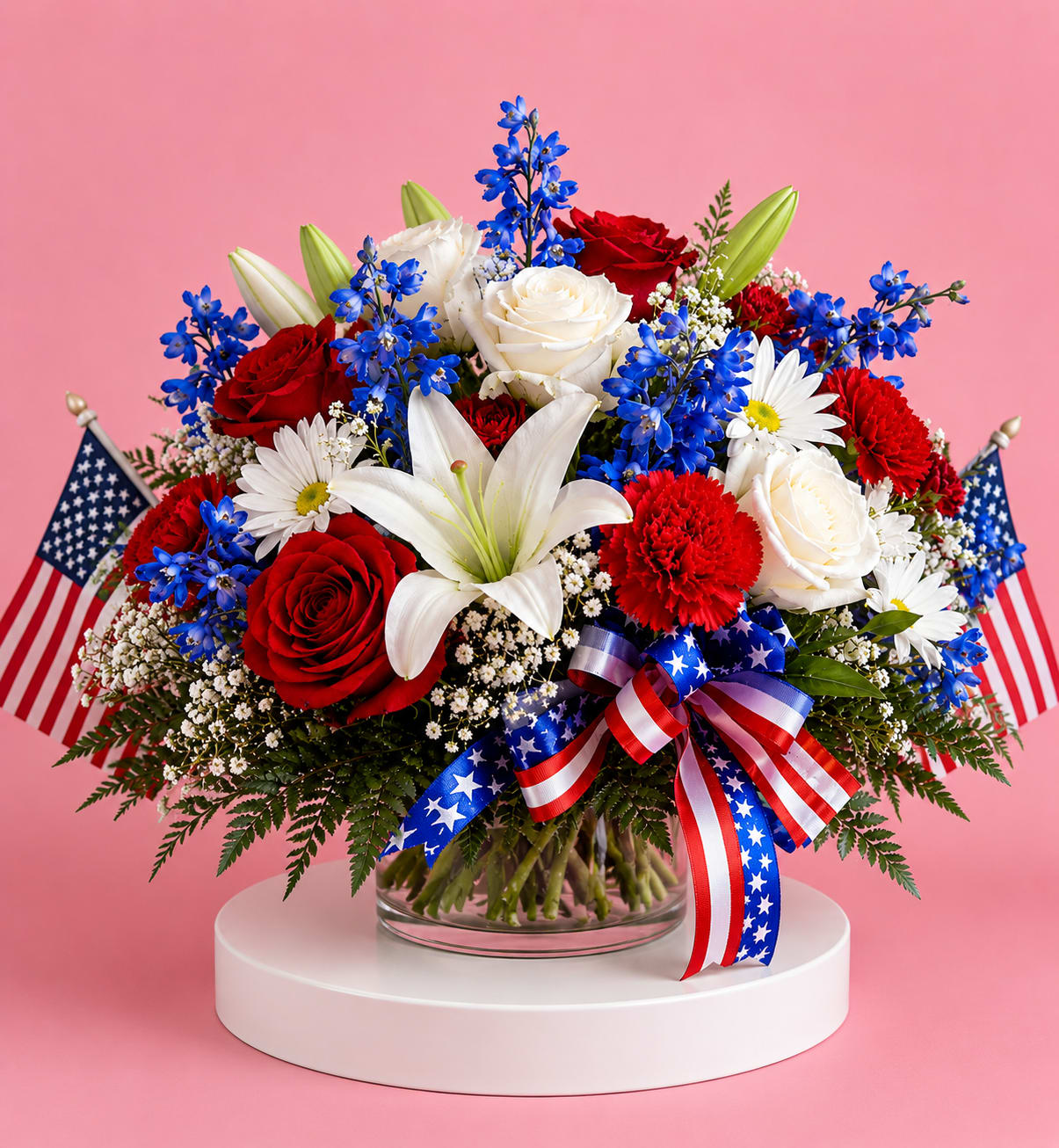 The Bristol 4th of July Bouquet - Red, white &amp; blue, roses, lilies, daisies and carnations in a clear cylinder vase with patriotic bows, small flags. A festive centerpiece for Fourth of July and summer celebrations.